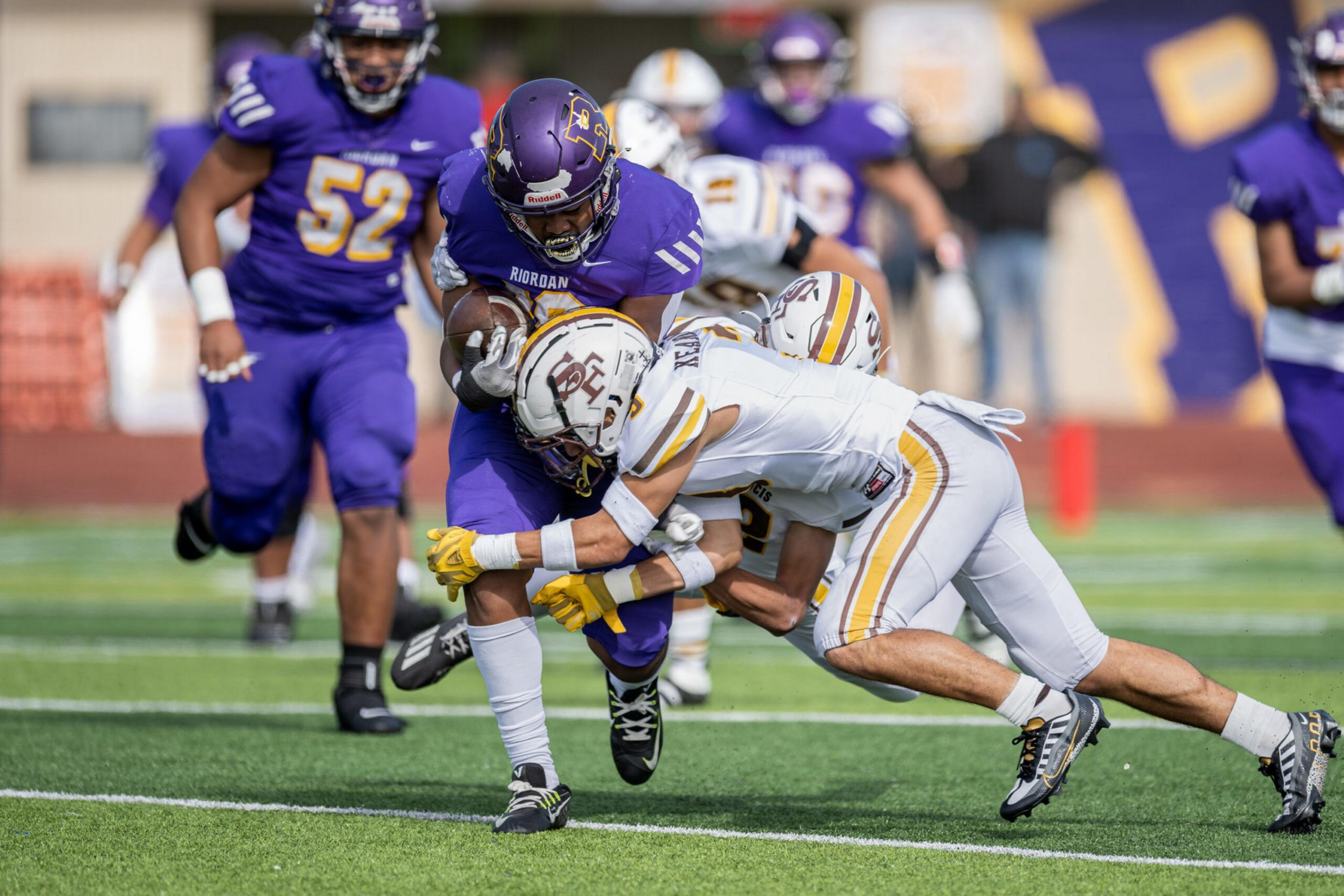 A player in purple grabs the football tightly while two players in white tackle him during an intense football game on a green field.