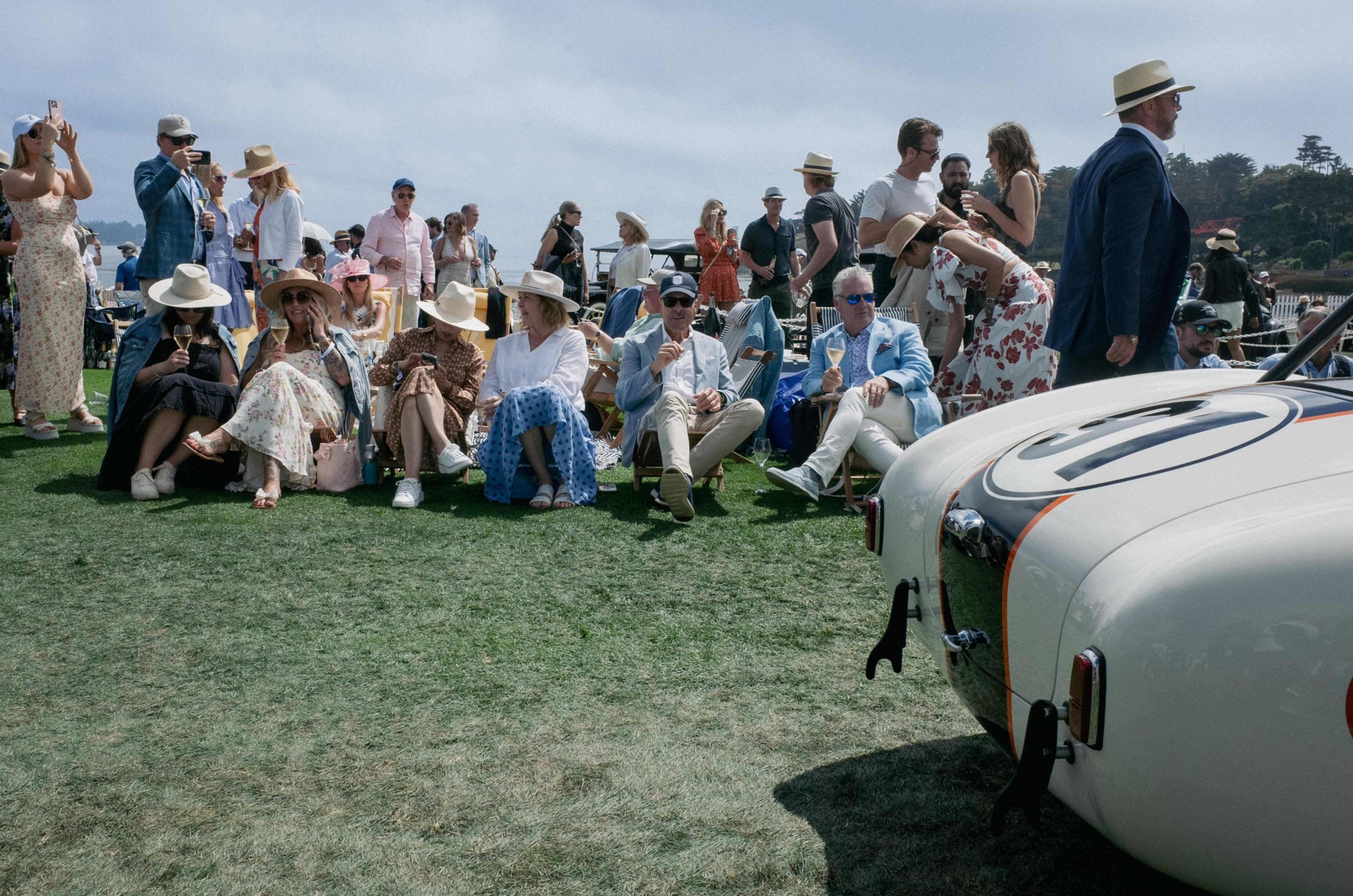 A group of well-dressed people sit and stand on grass, many wearing hats, near a white vintage car with racing decals on a sunny day.