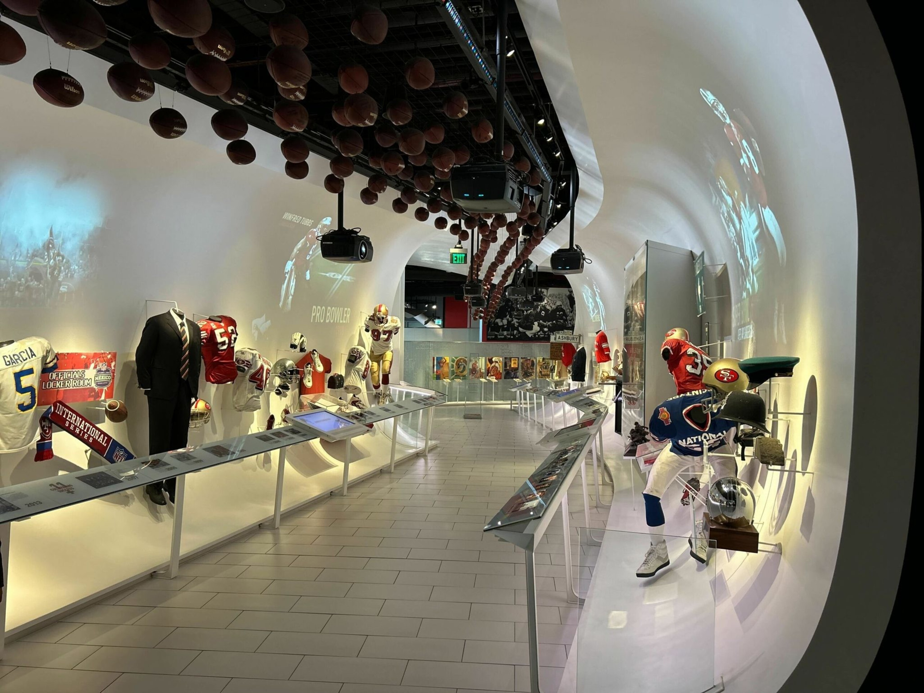 Football jerseys, helmets, and memorabilia are displayed along curved white walls with suspended footballs overhead and projectors showing player images.