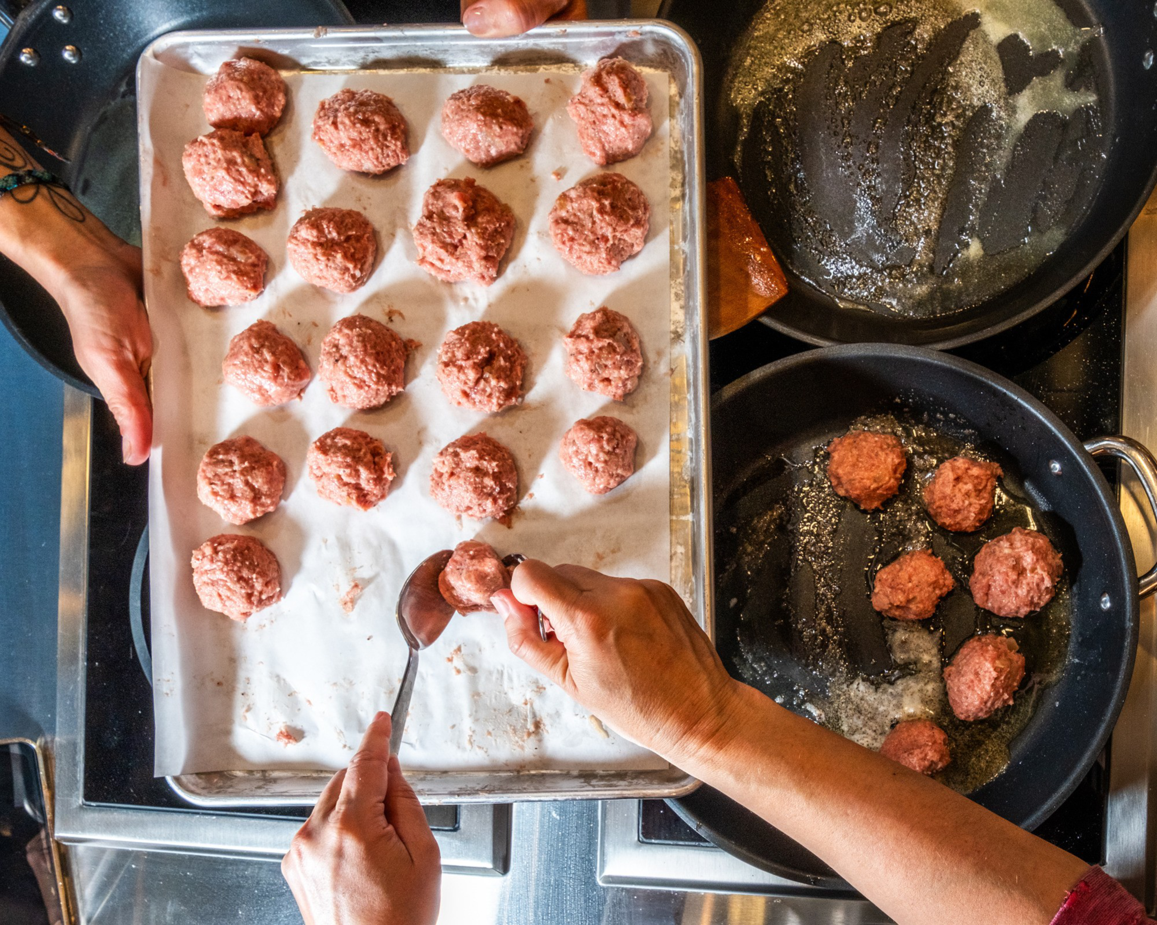 Two hands are shaping meatballs on a tray, while several cook in a buttered pan. The scene is an overhead view of a busy kitchen setup.