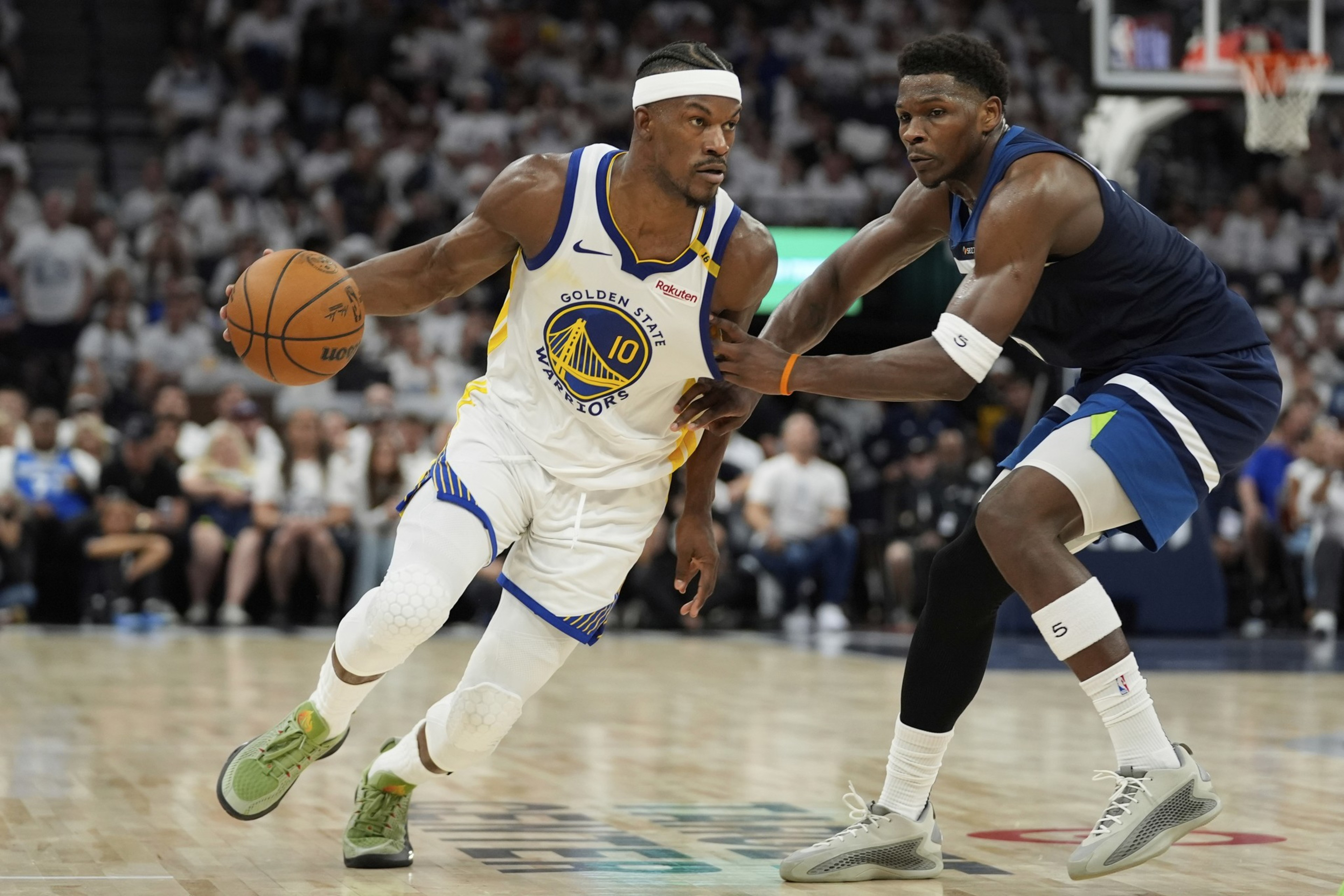 Two basketball players in action on the court, one dribbling the ball wearing a white Golden State Warriors jersey, the other defending in a blue uniform.