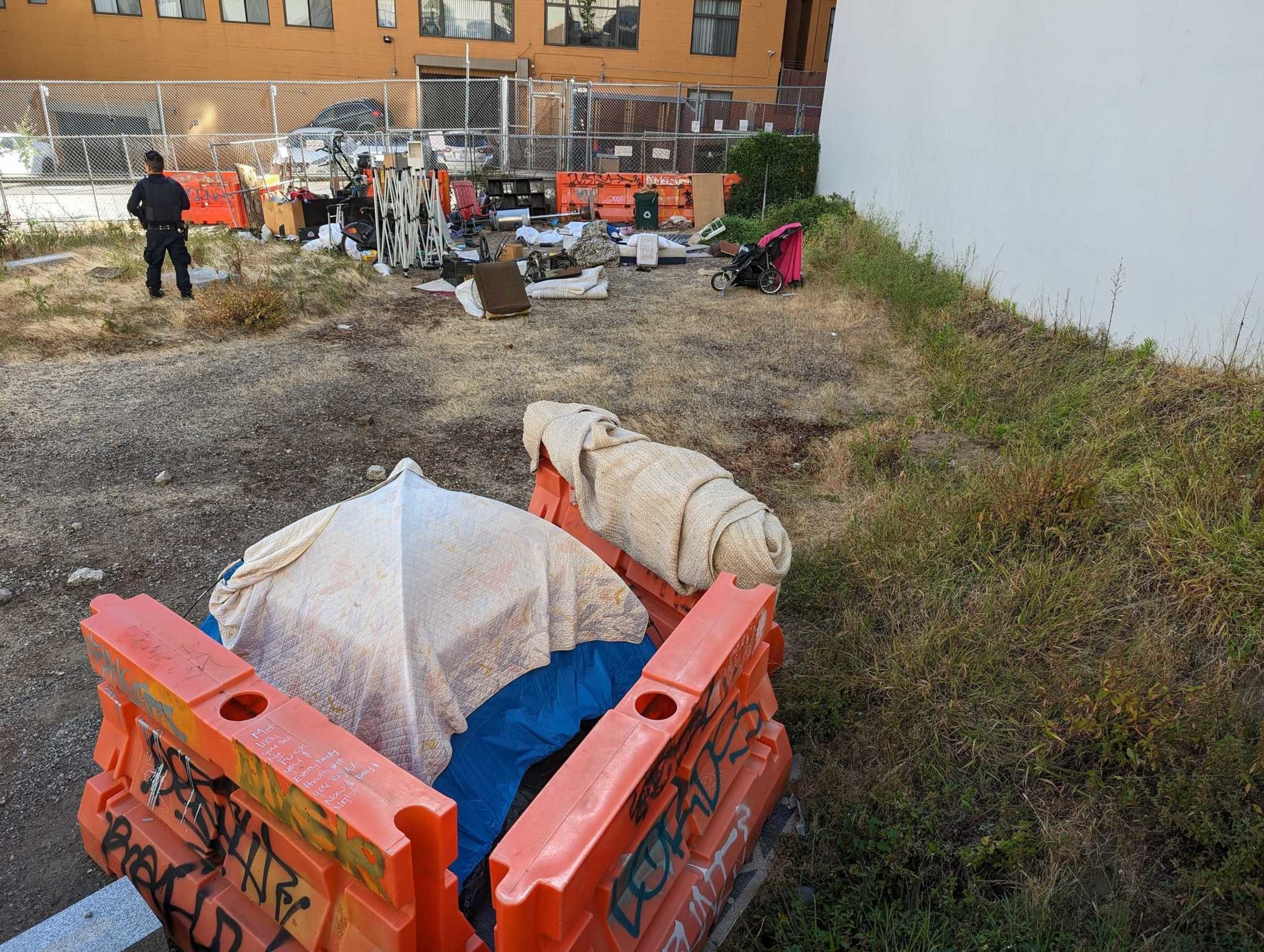 A person stands by a fence amidst scattered belongings and debris, including a stroller and barricades in a grassy, vacant lot near a building.