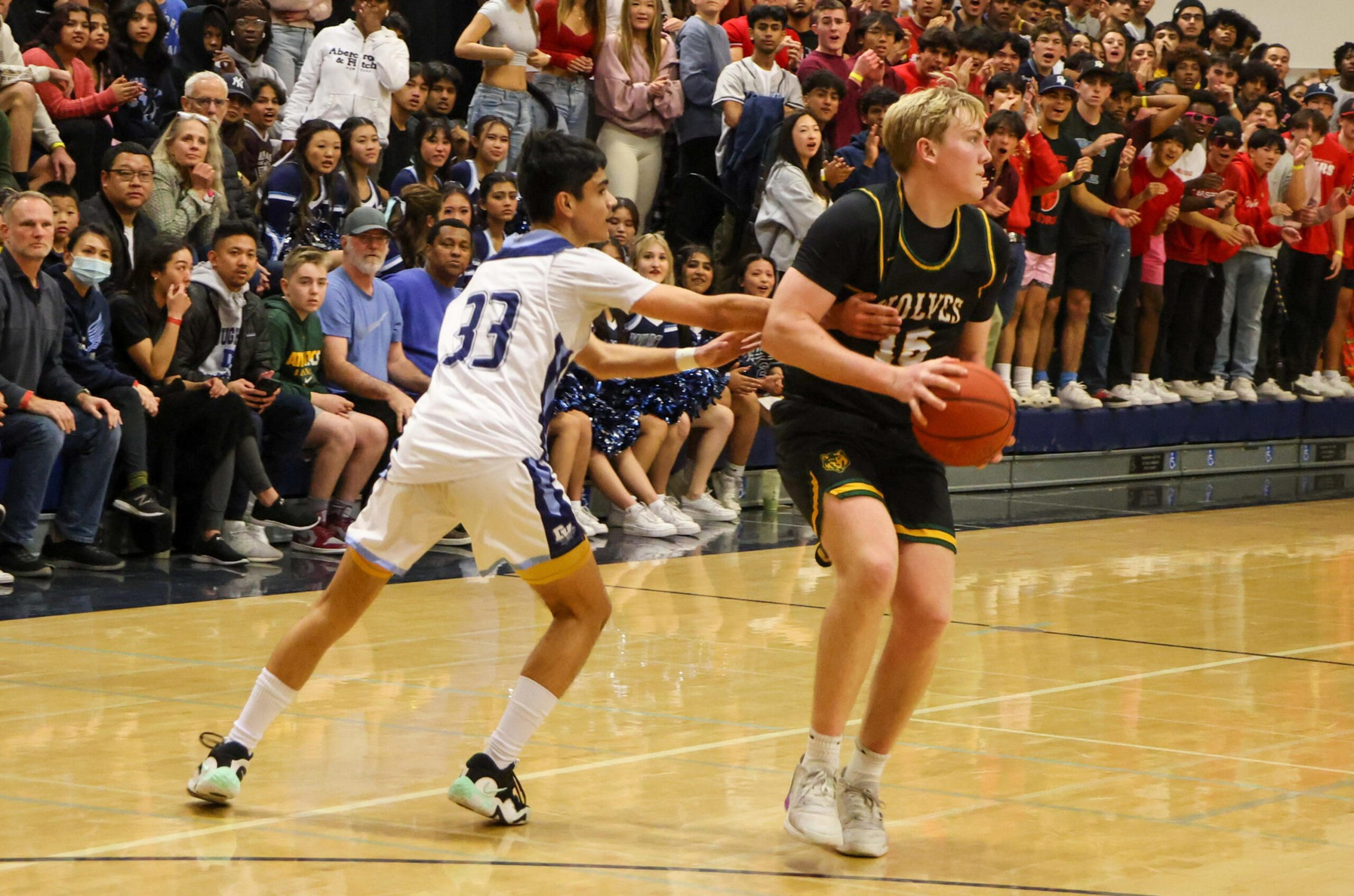 A basketball player in a black Wolves uniform holds the ball while being closely defended by a player in a white jersey, with a crowded gym audience watching.