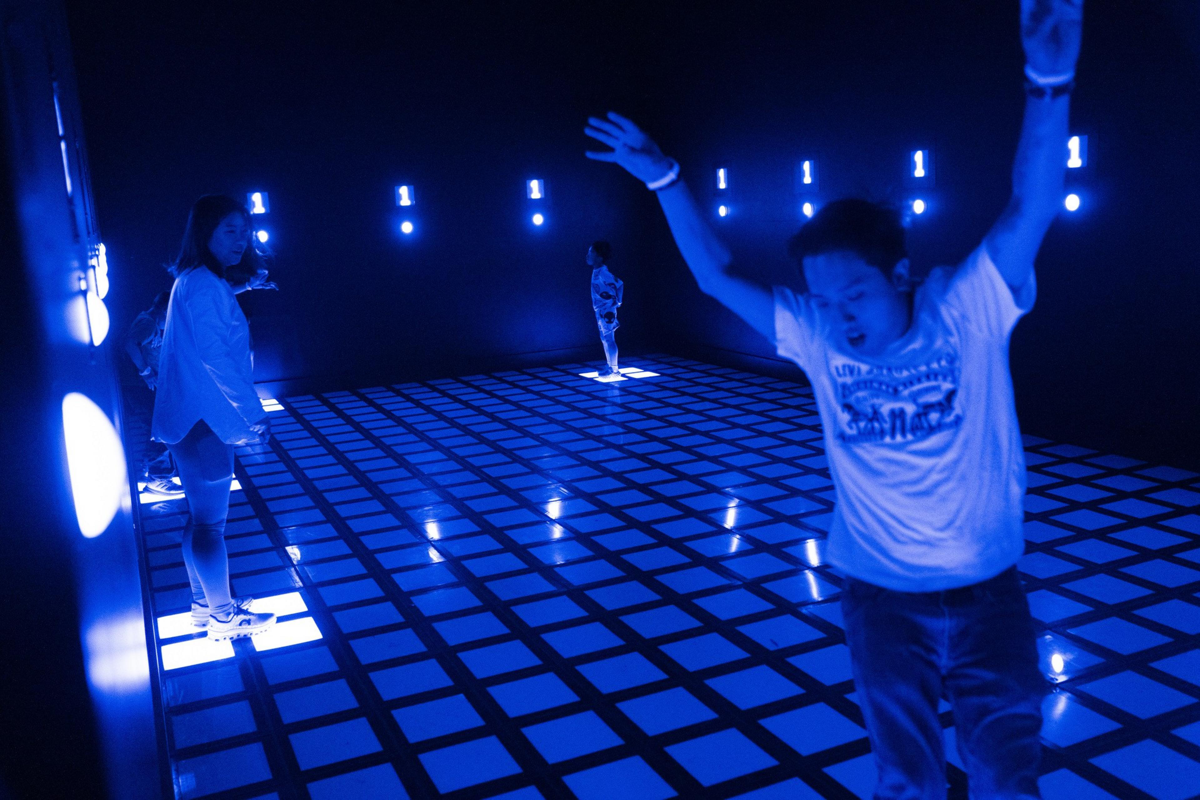 People are interacting in a dark room with a glowing blue grid floor, illuminated by blue lights on the walls and some individuals standing on lit squares.