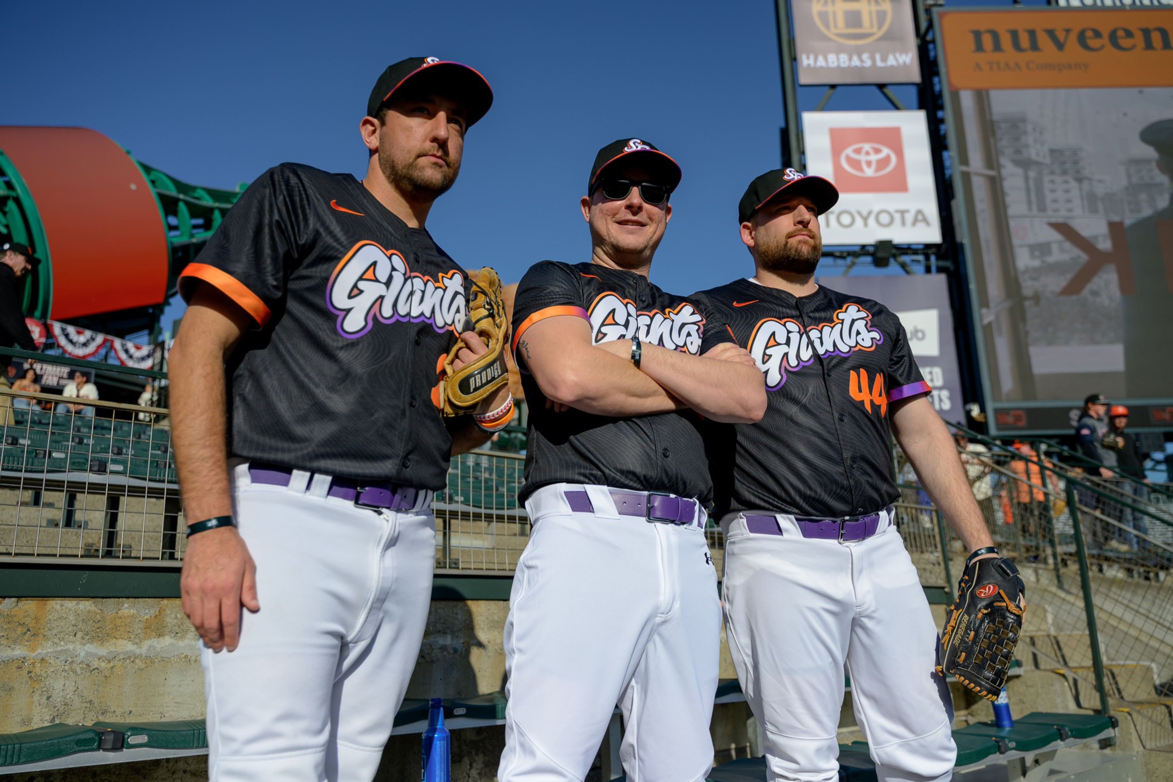Three baseball players stand confidently, wearing black jerseys with &quot;Giants&quot; in colorful letters, white pants, and caps. Two are holding gloves, with a blue sky behind.