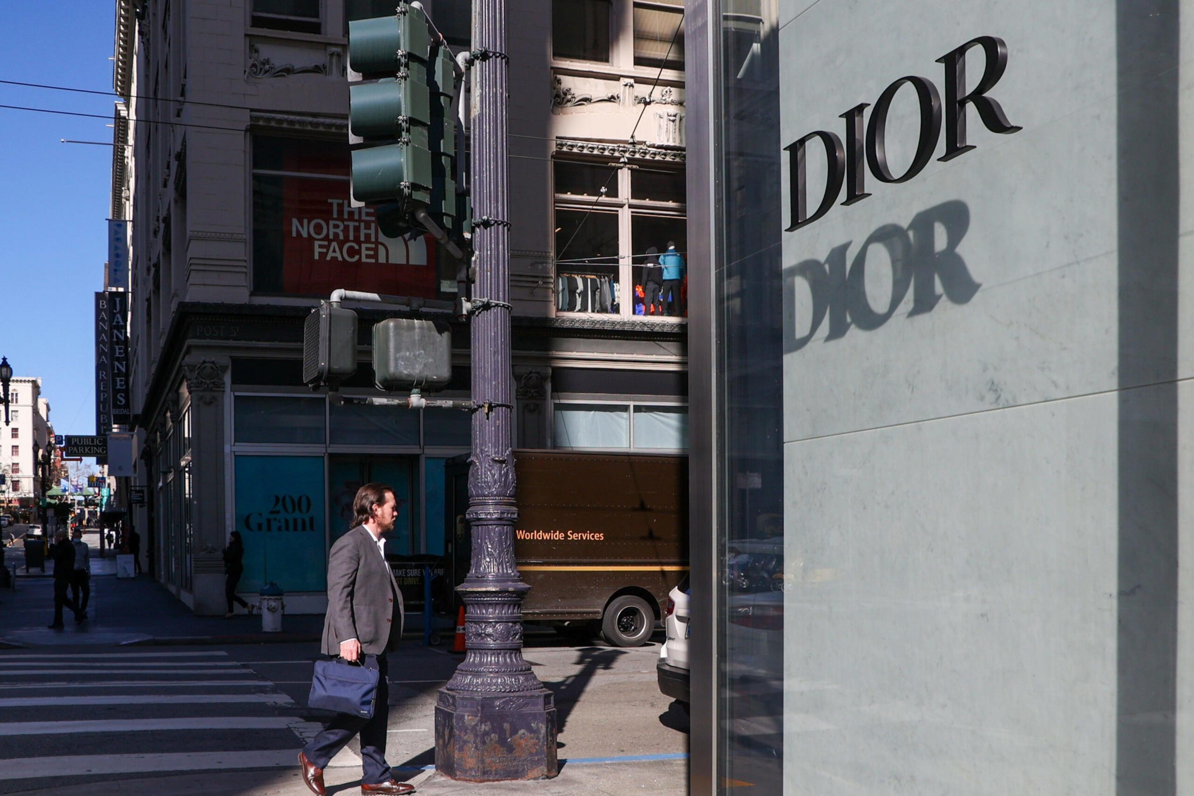 A man in business attire crosses a street at an intersection near a tall lamp post and a Dior store with its logo casting a shadow.