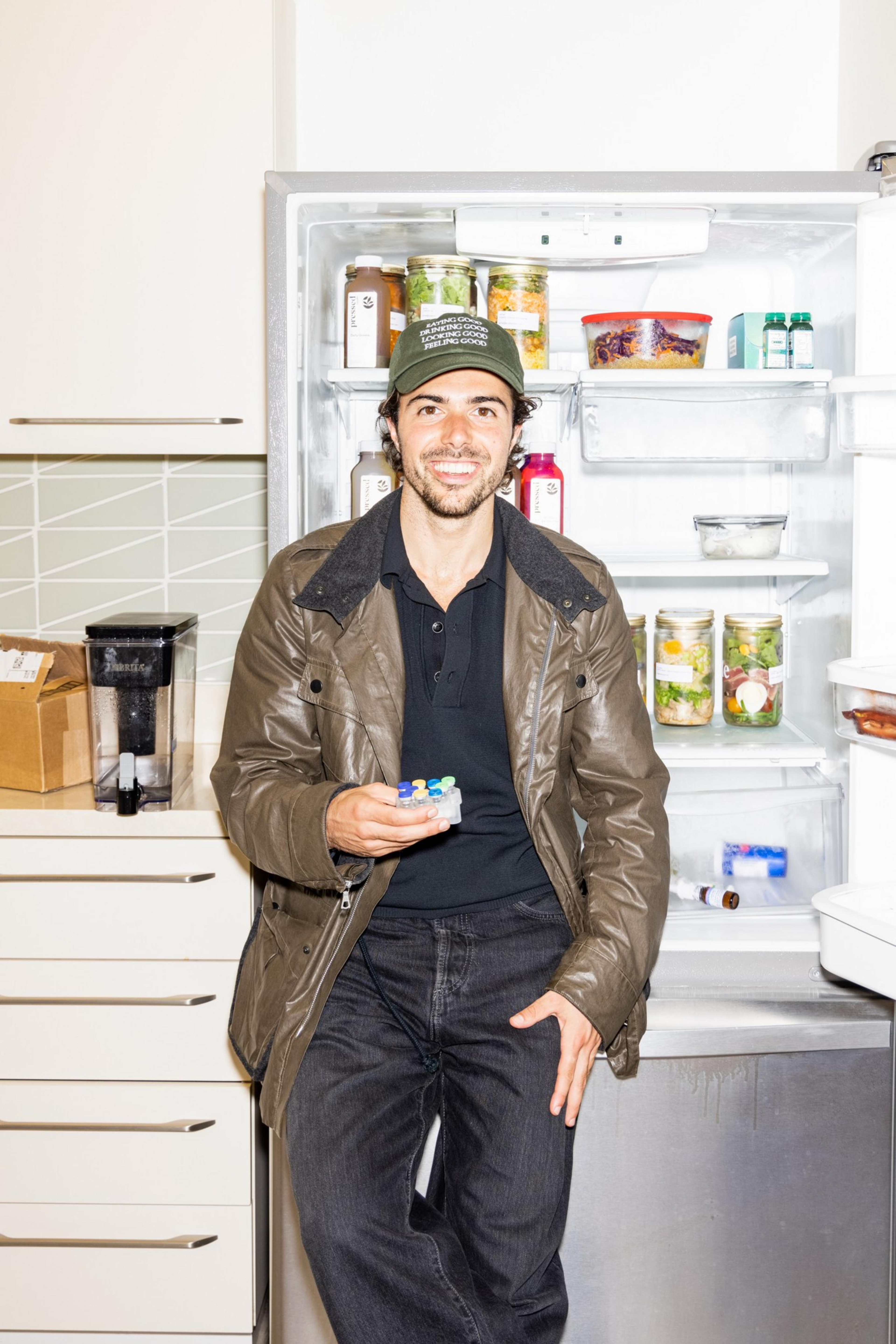 A smiling man in a brown jacket and green hat stands by an open refrigerator, holding a pack of colorful pills in a modern kitchen.