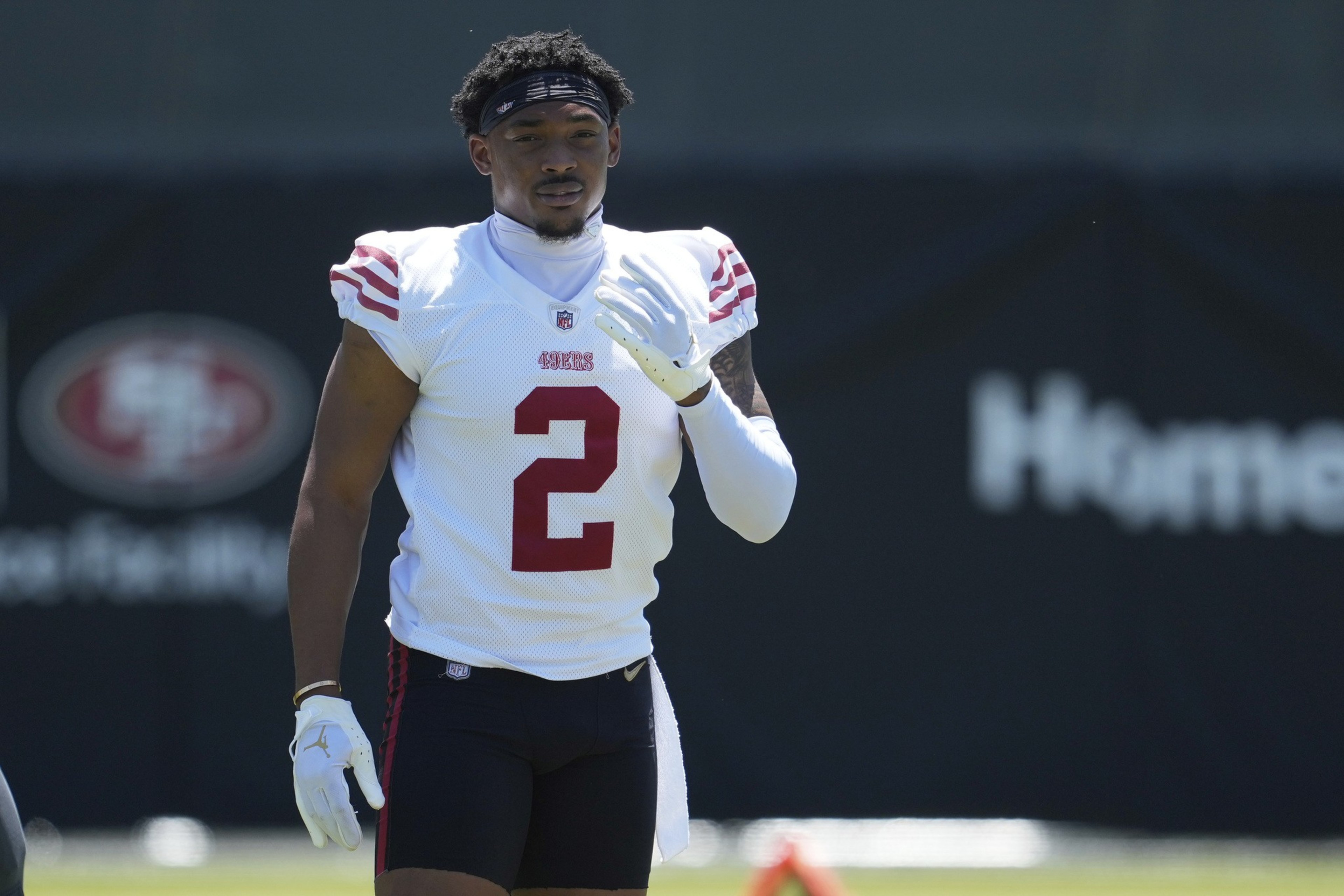 A football player in a white jersey with red stripes and the number 2 is on a field. He wears white gloves and a dark headband, walking towards the camera.