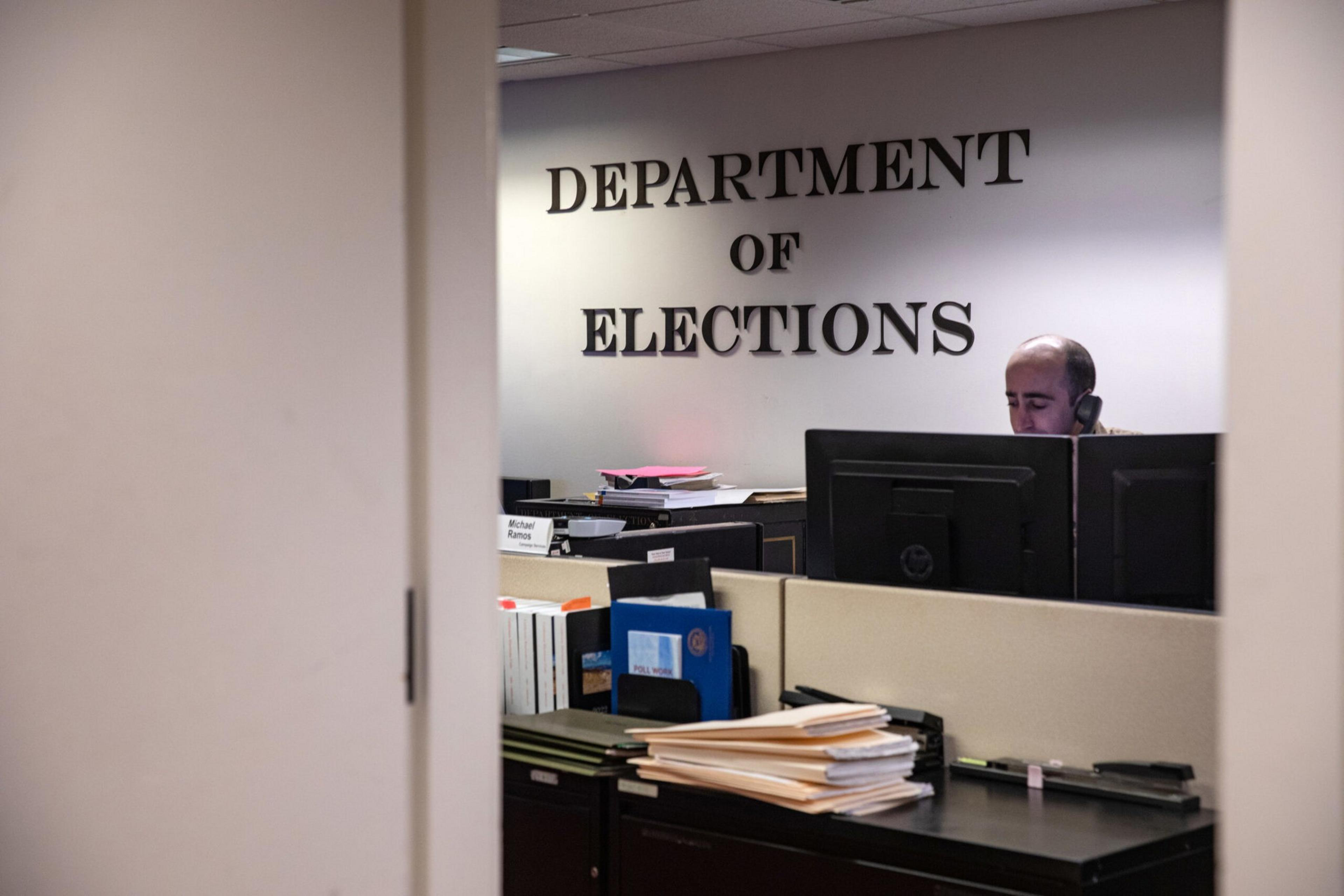 A man is sitting at a desk with two computer monitors inside an office labeled "Department of Elections," surrounded by files and folders.