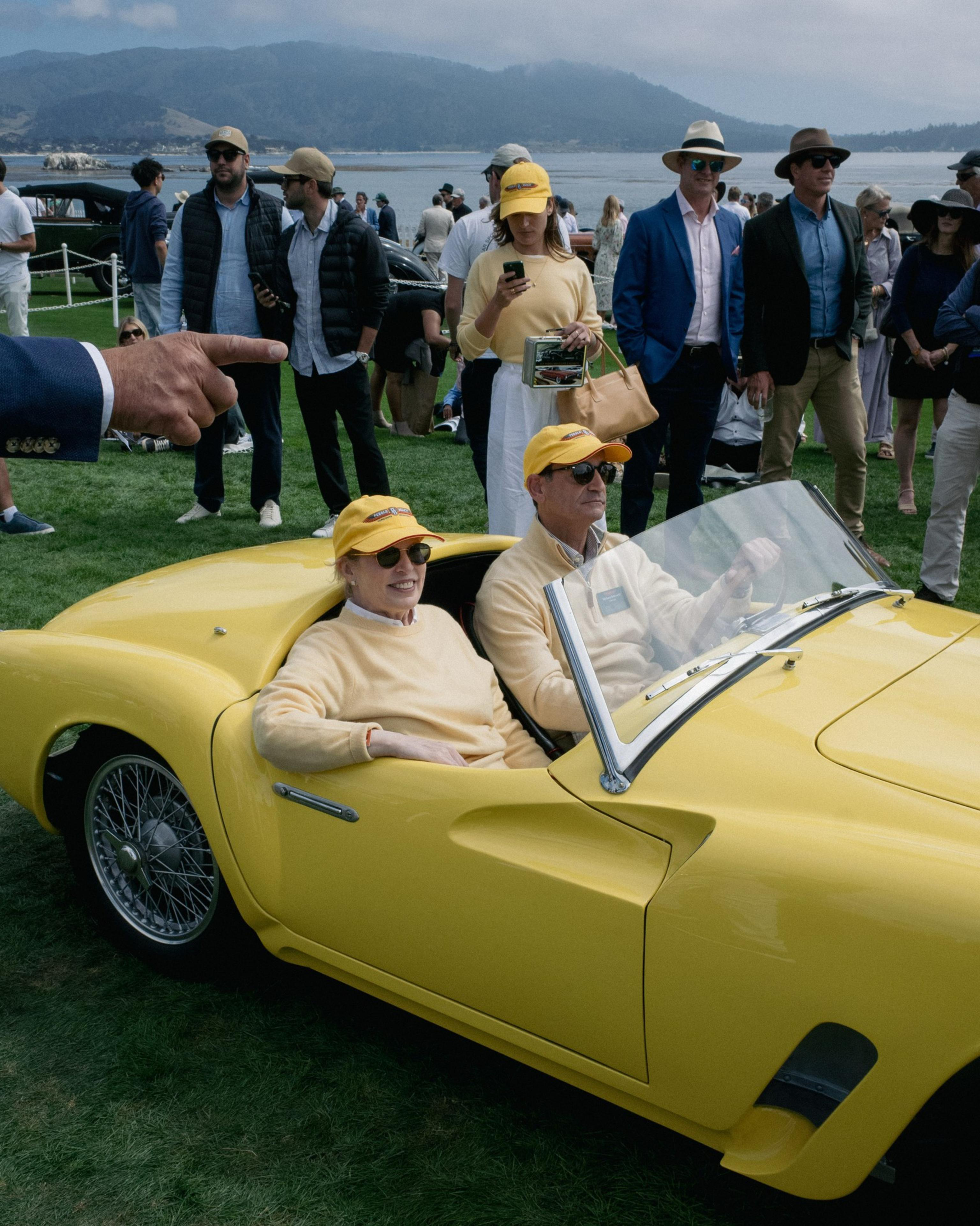 Two people wearing matching yellow hats and light sweaters sit in a bright yellow vintage convertible car on a lawn, surrounded by several casually dressed onlookers.