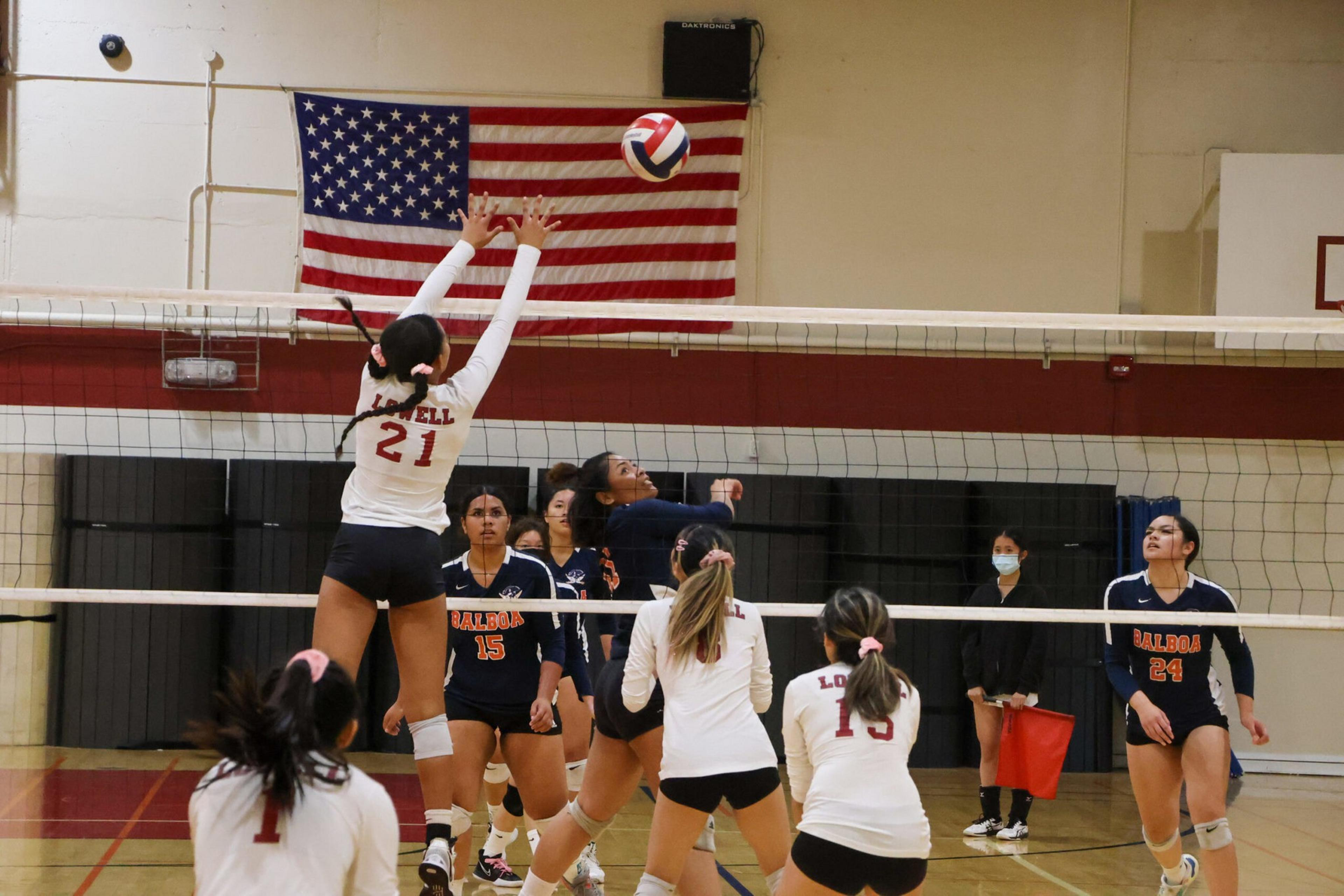 A volleyball player in a white jersey jumps to block a ball while players from both teams watch in a gym with an American flag on the wall.
