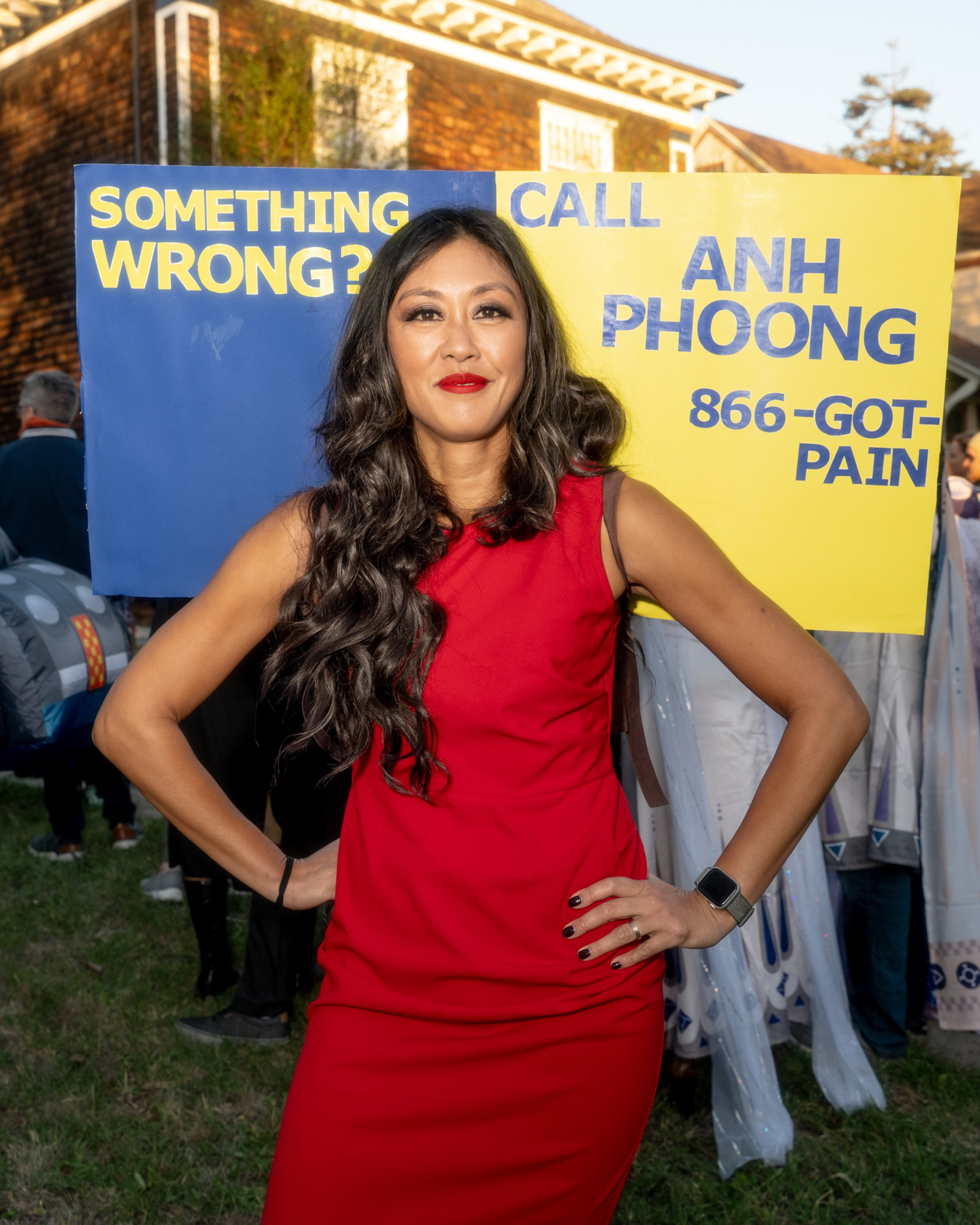 A woman in a red dress poses confidently in front of a sign that reads, “Something wrong? Call Anh Phoong 866-GOT-PAIN.”