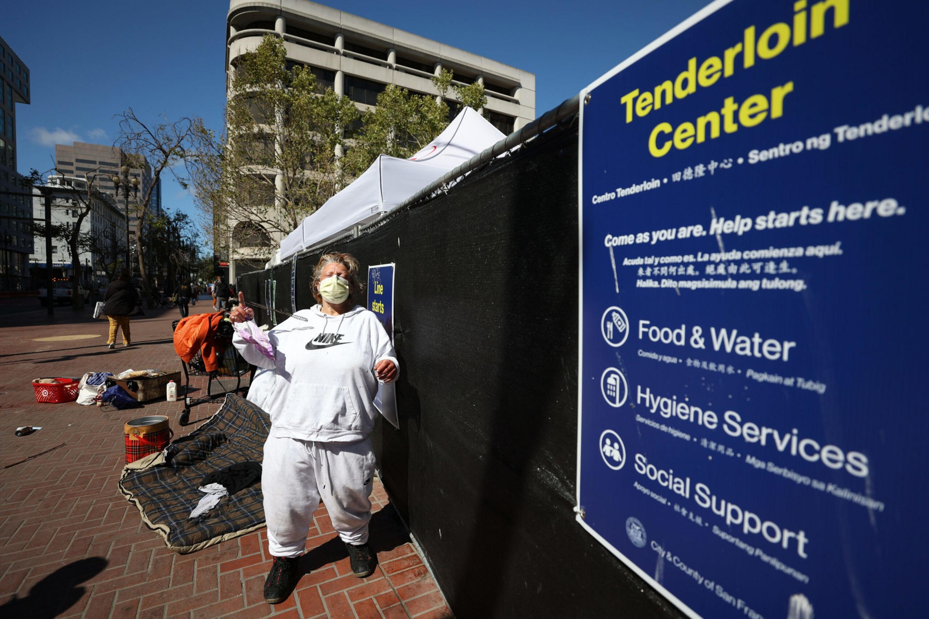 A person in white stands by a sign for a support center on a city street, with tents and belongings nearby.