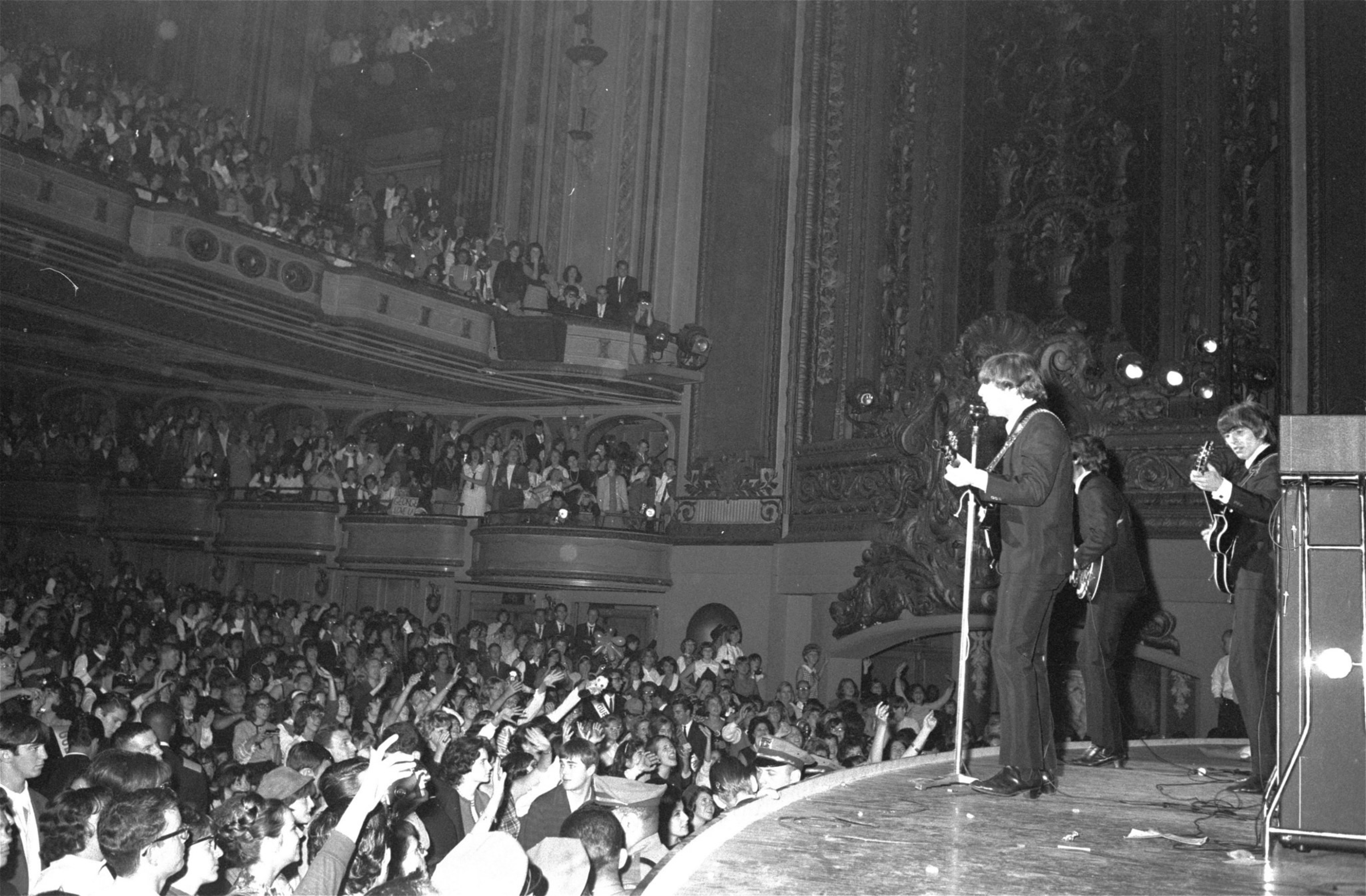 A black and white photo of a band performing on stage with a lively and packed audience in an ornate theater.