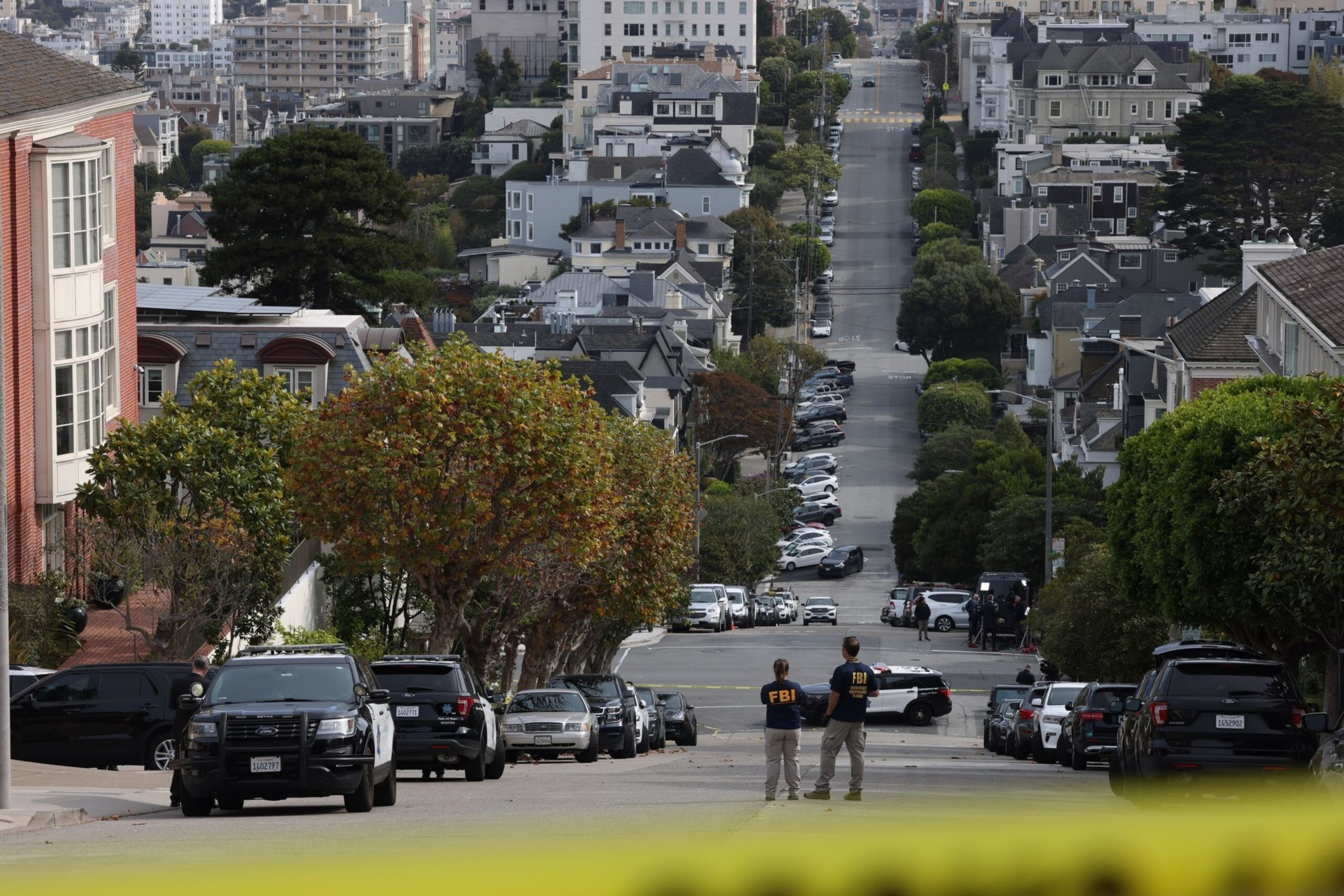 Two FBI agents stand on a steep residential street lined with parked cars and trees, with a cityscape of houses and buildings in the background.