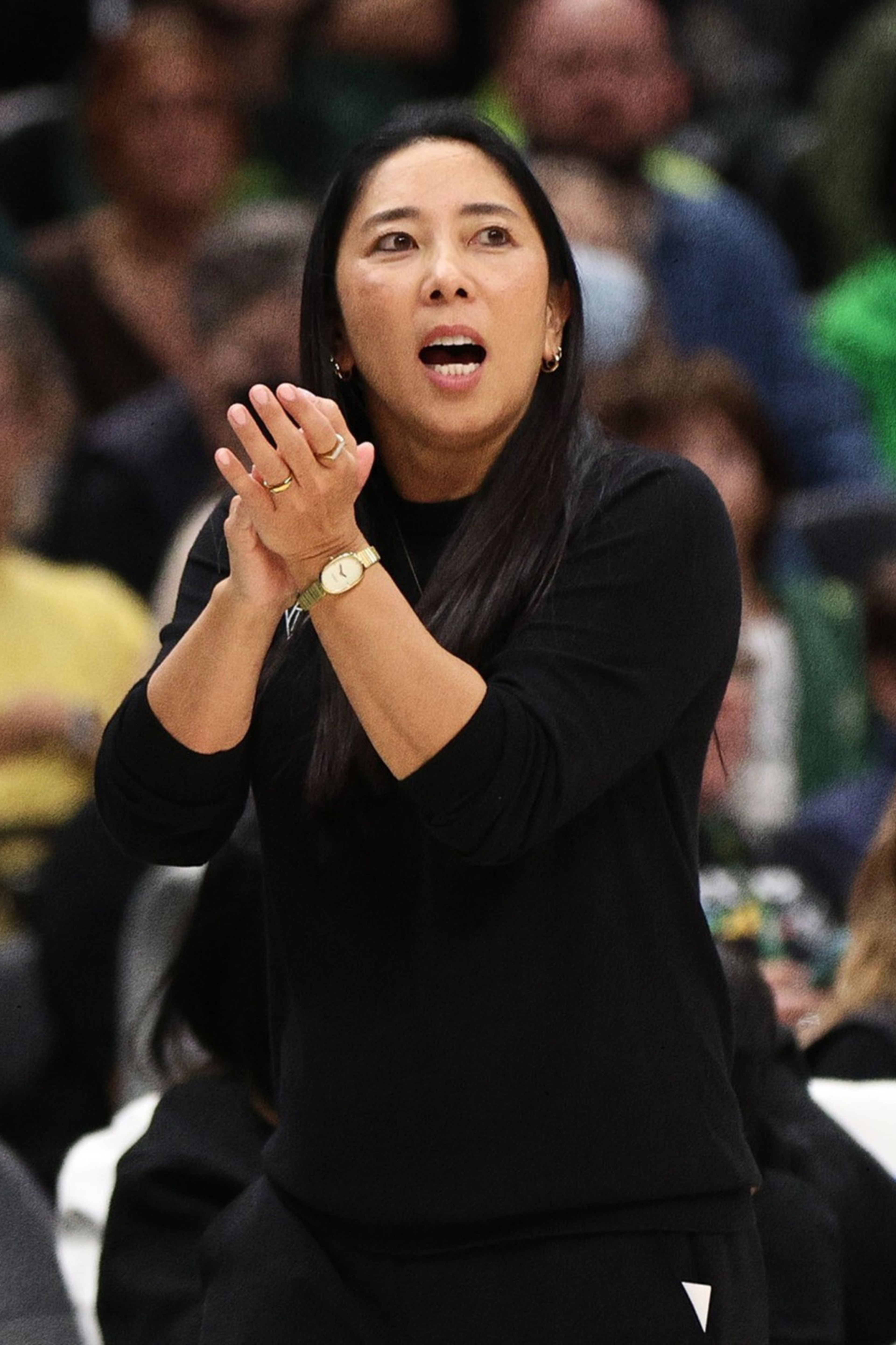 A woman in black claps her hands and speaks, standing in a crowded arena with a basketball-themed red and black graphic on the left side.