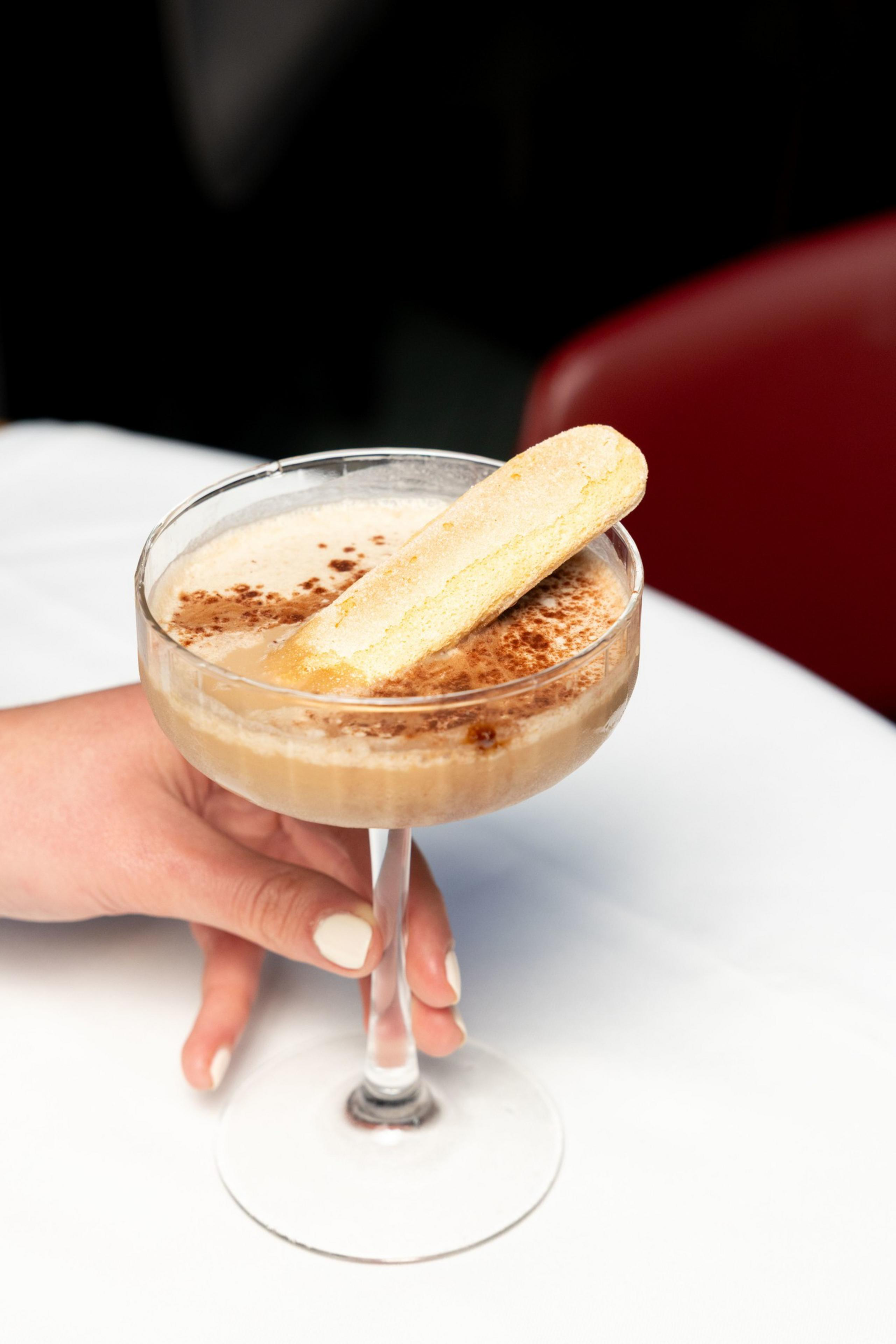 A hand holds a glass with a creamy drink topped with cocoa and a ladyfinger biscuit, set against a white background.