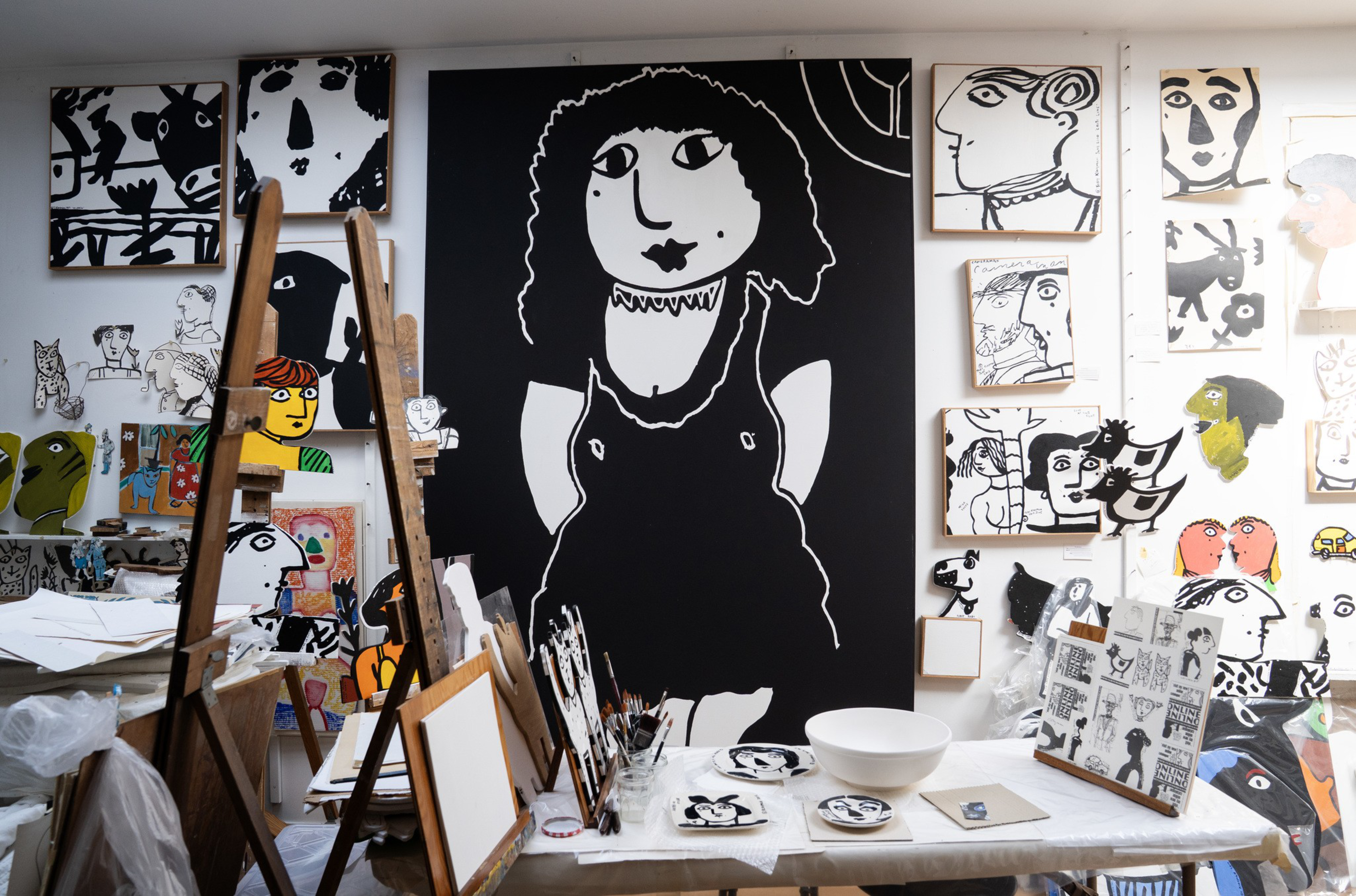 A studio filled with black-and-white and colorful abstract portraits, featuring a large black canvas with a white cartoonish woman in the center.