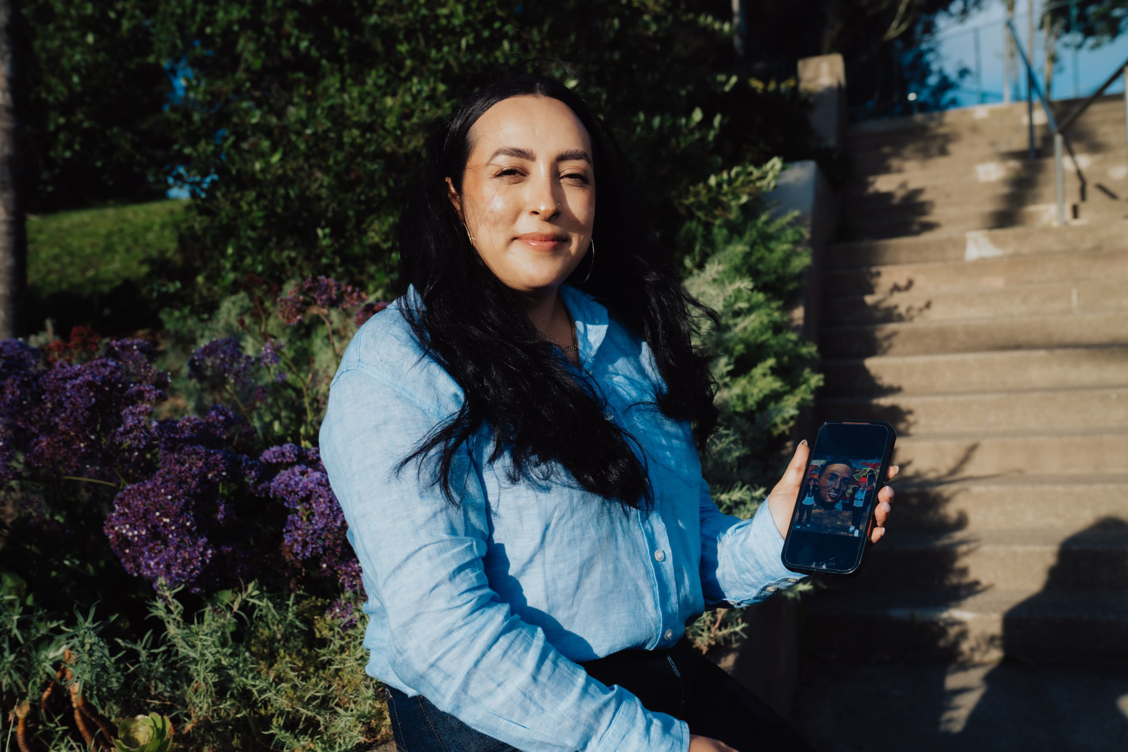 A woman with long dark hair and a light blue shirt sits near purple flowers, smiling and holding a phone showing a selfie.