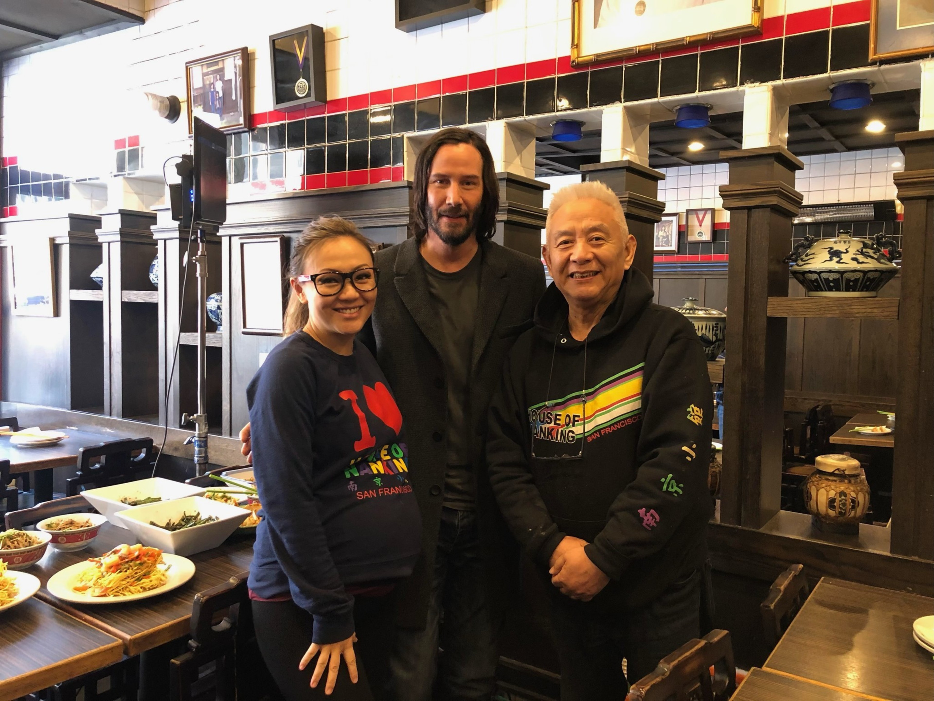 Three people stand smiling in a restaurant dining area with plates of food on tables, including a pregnant woman wearing glasses and a sweatshirt.