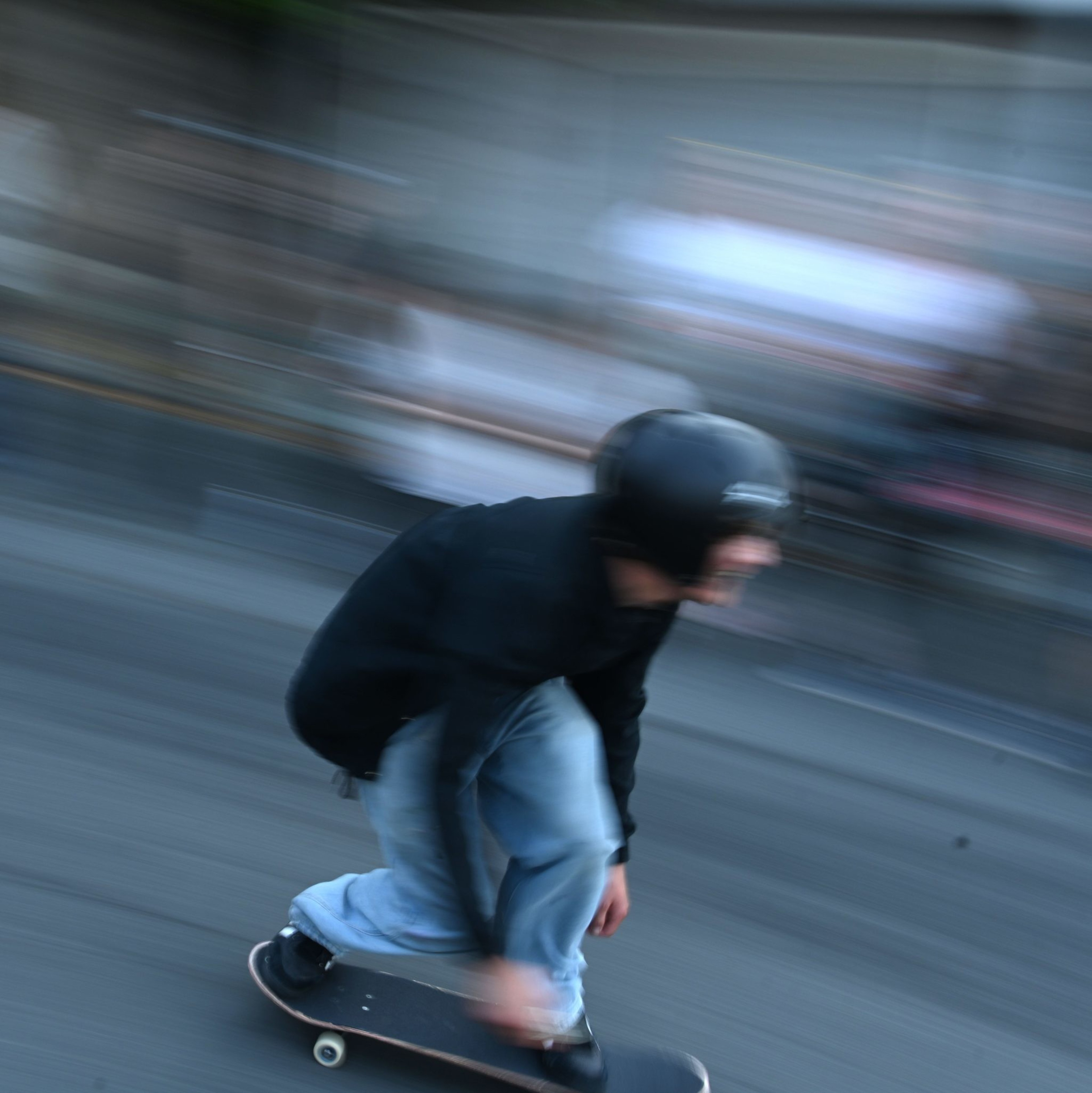 A person wearing a helmet and dark clothing is skateboarding rapidly on a street, with a blurred background indicating high speed.