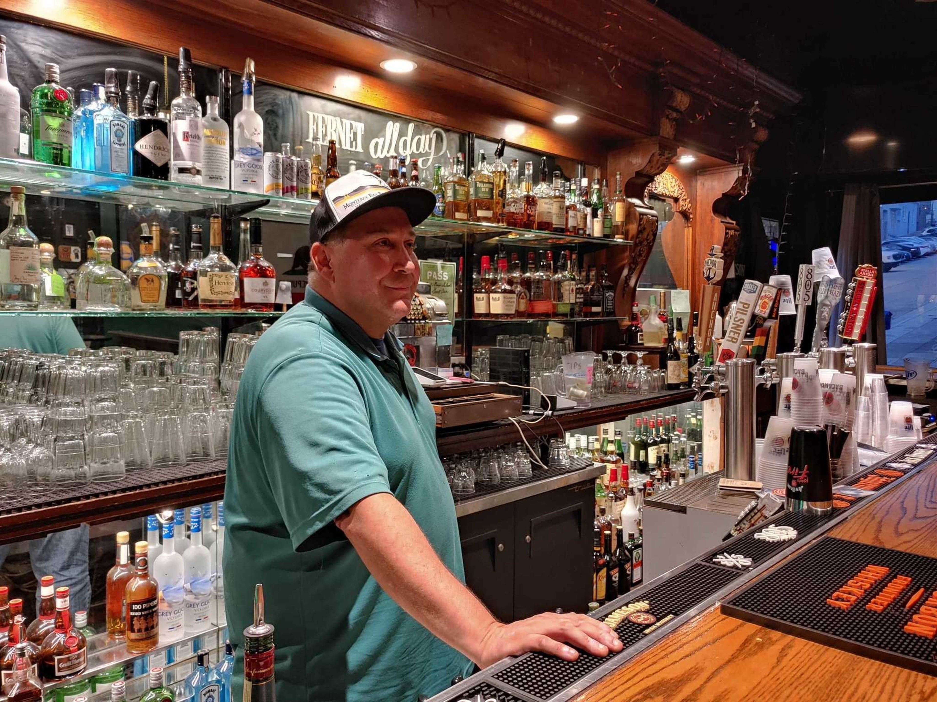 A man in a teal shirt and a cap stands behind a wooden bar filled with bottles, glasses, and multiple beer taps.