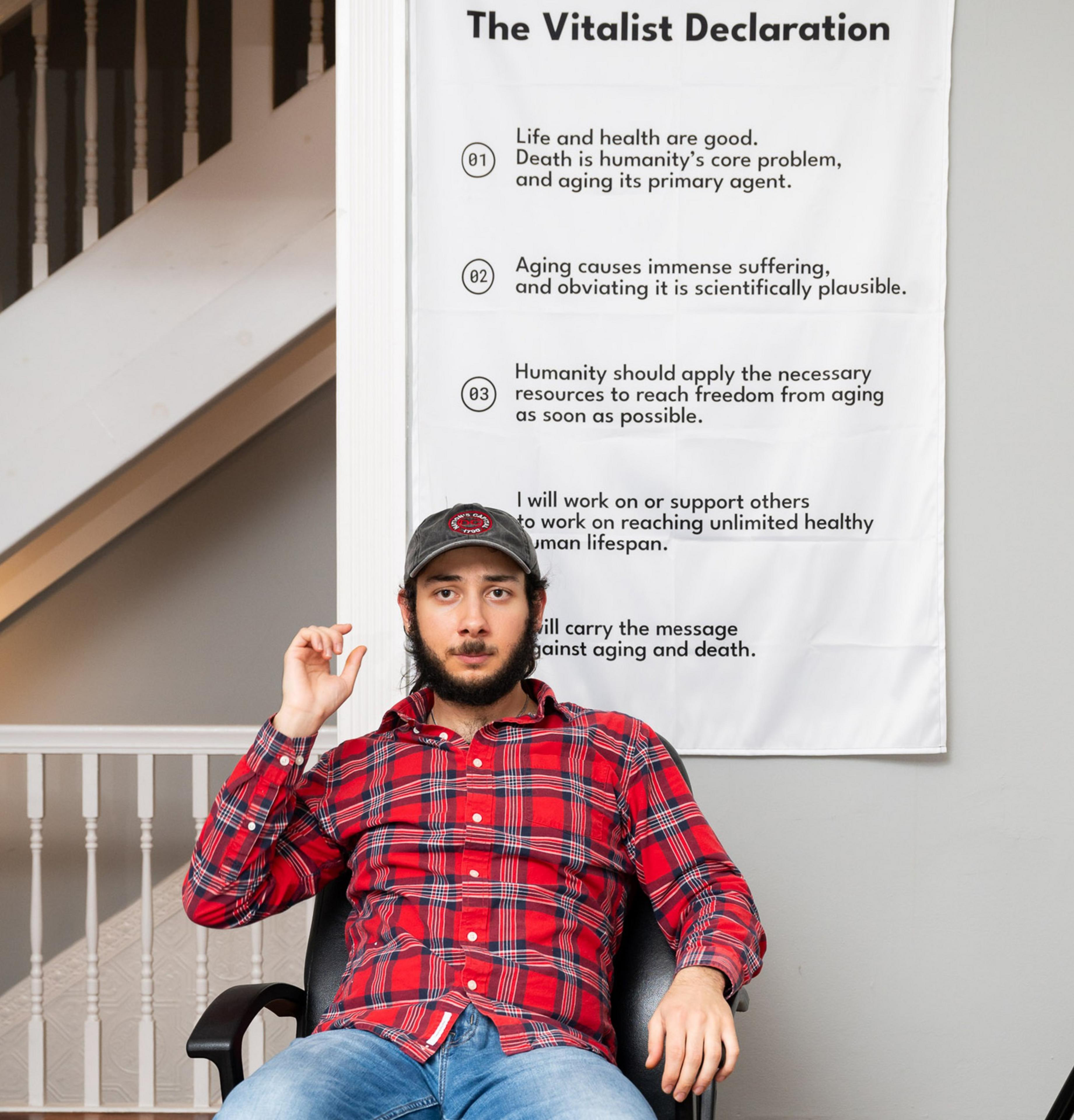 A man in a plaid shirt and cap sits in front of a poster titled &quot;The Vitalist Declaration.&quot; The text discusses life, health, aging, and death.