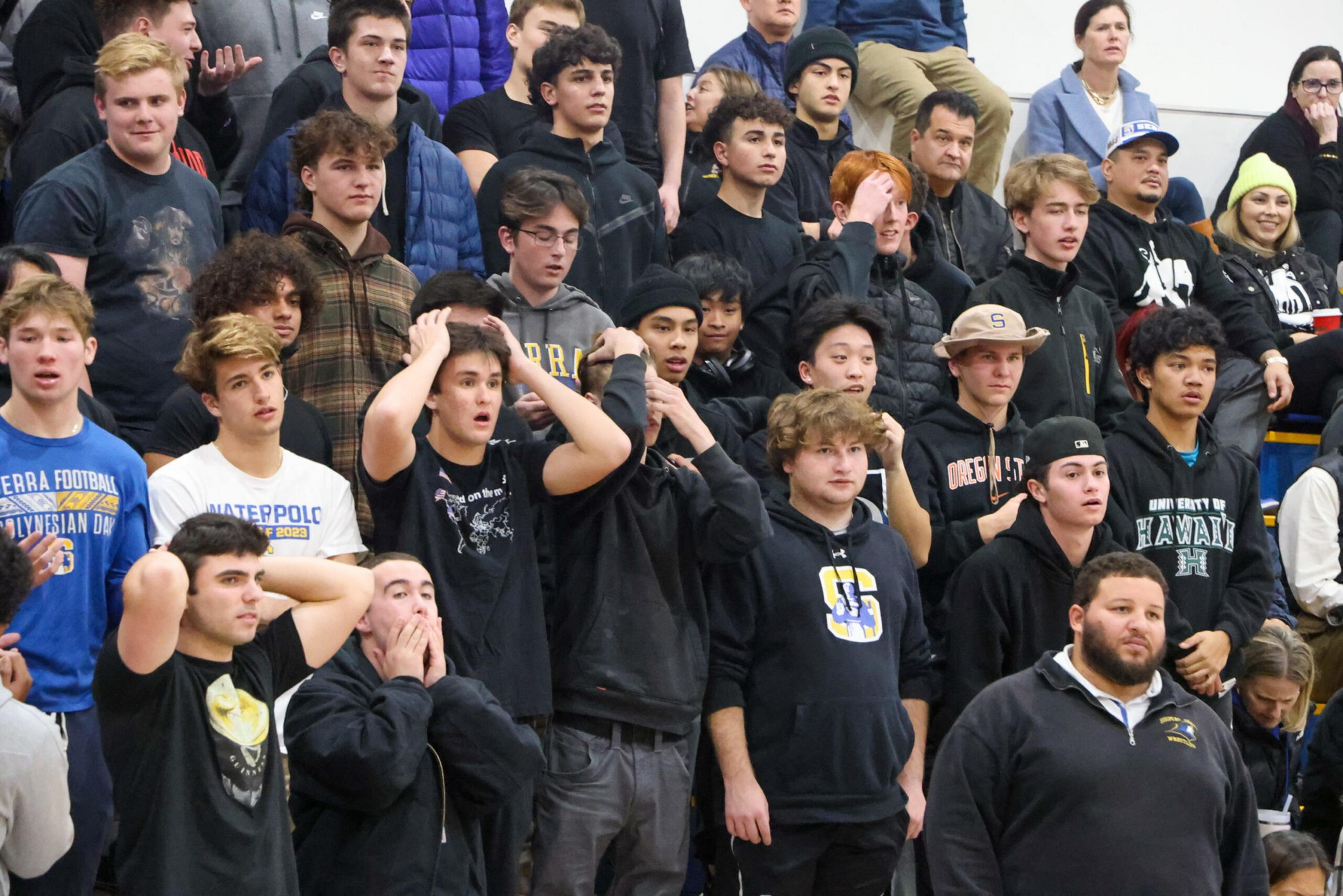A crowd of mostly young men and boys, some with surprised or anxious expressions, stand closely together, many wearing casual hoodies and jackets.