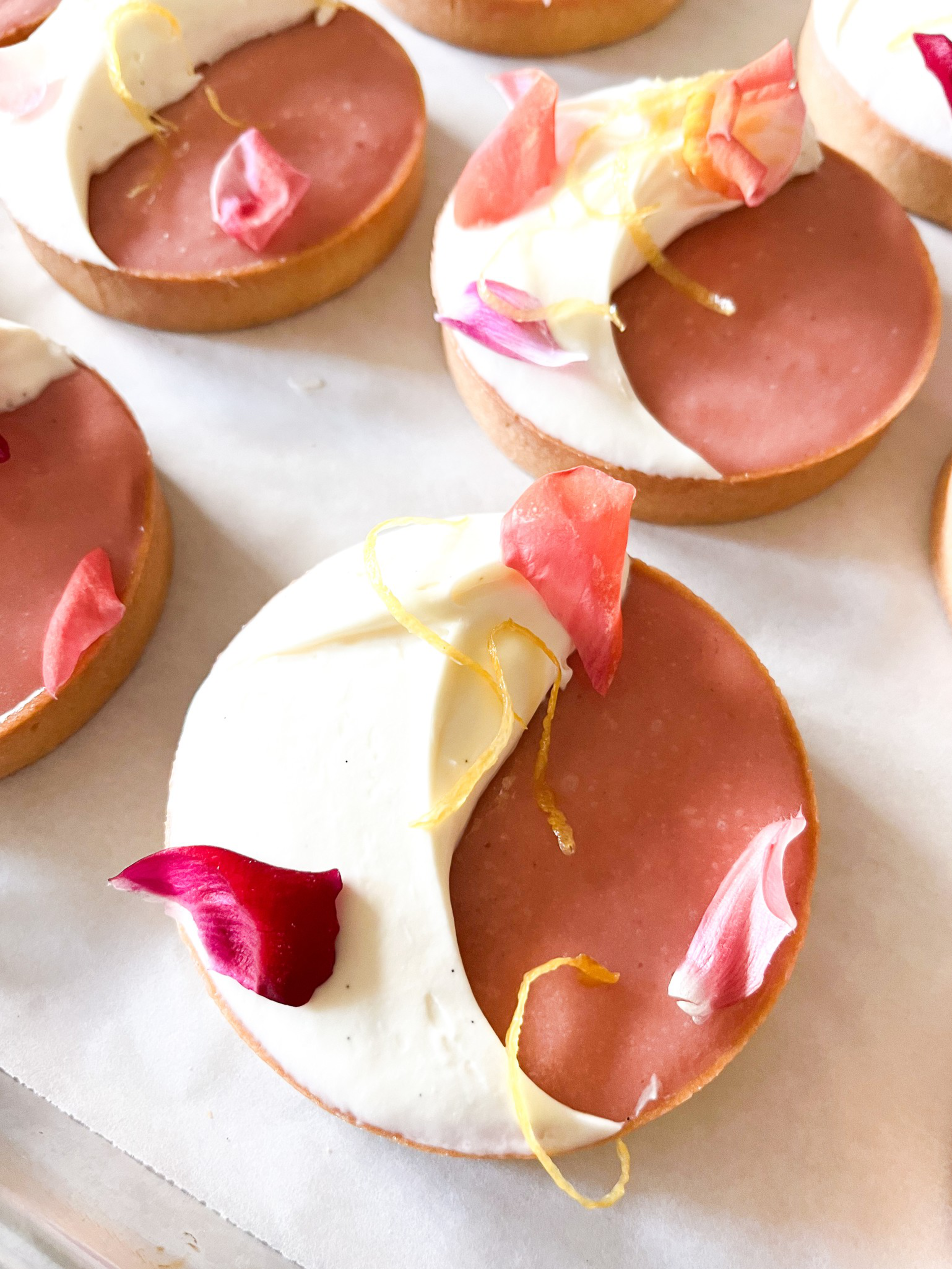 Round tartlets with half covered in smooth cream and half in a pink glaze, decorated with colorful edible flower petals and thin orange zest strands.