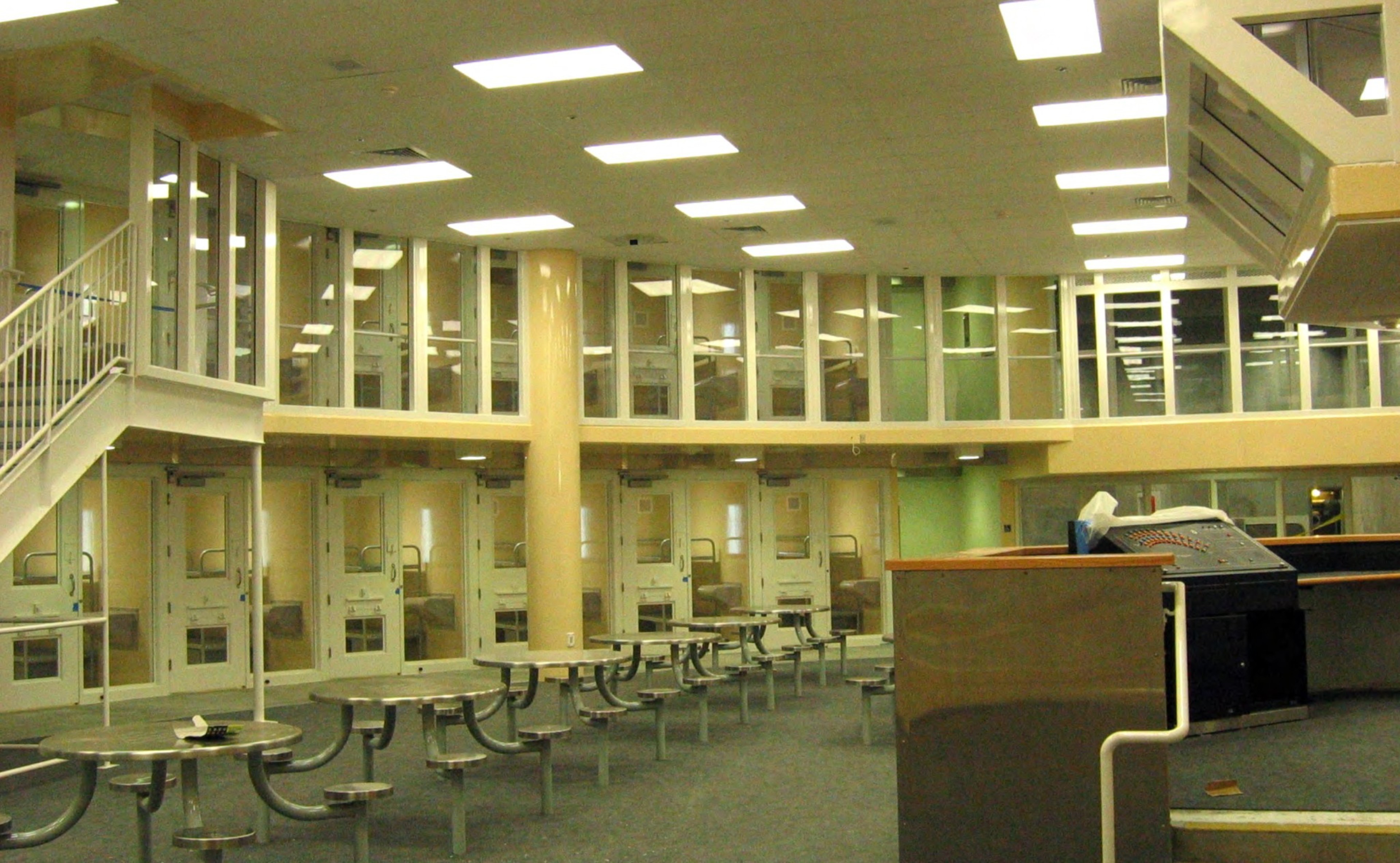 The image shows an institutional setting with metal tables and benches, two levels of glass-fronted rooms, and a control desk in the foreground under bright lighting.