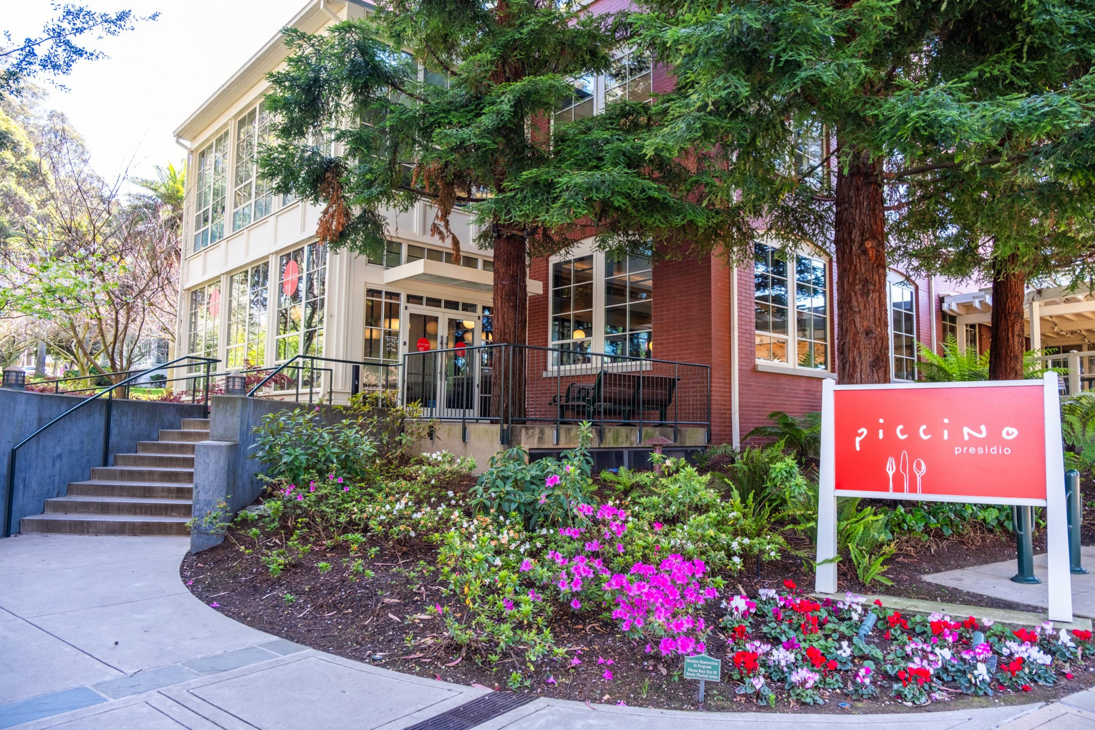 The image shows a building with large windows surrounded by lush greenery and colorful flowers. A red sign in front reads &quot;Piccino Presidio.&quot;