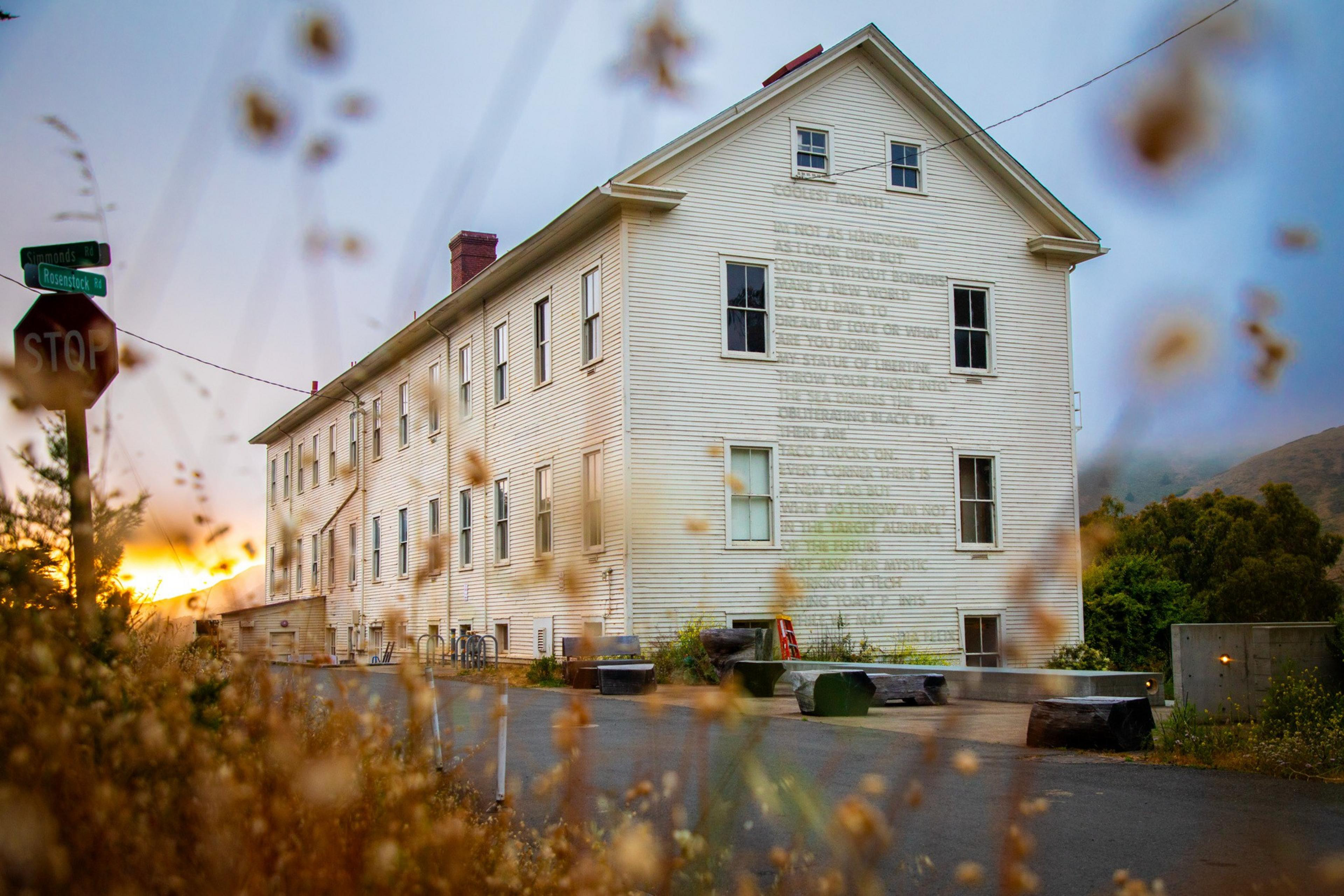 An old, white wooden building with many windows stands against a hazy sky. Blurry wildflowers frame the foreground, with a sunset peeking through on the left.