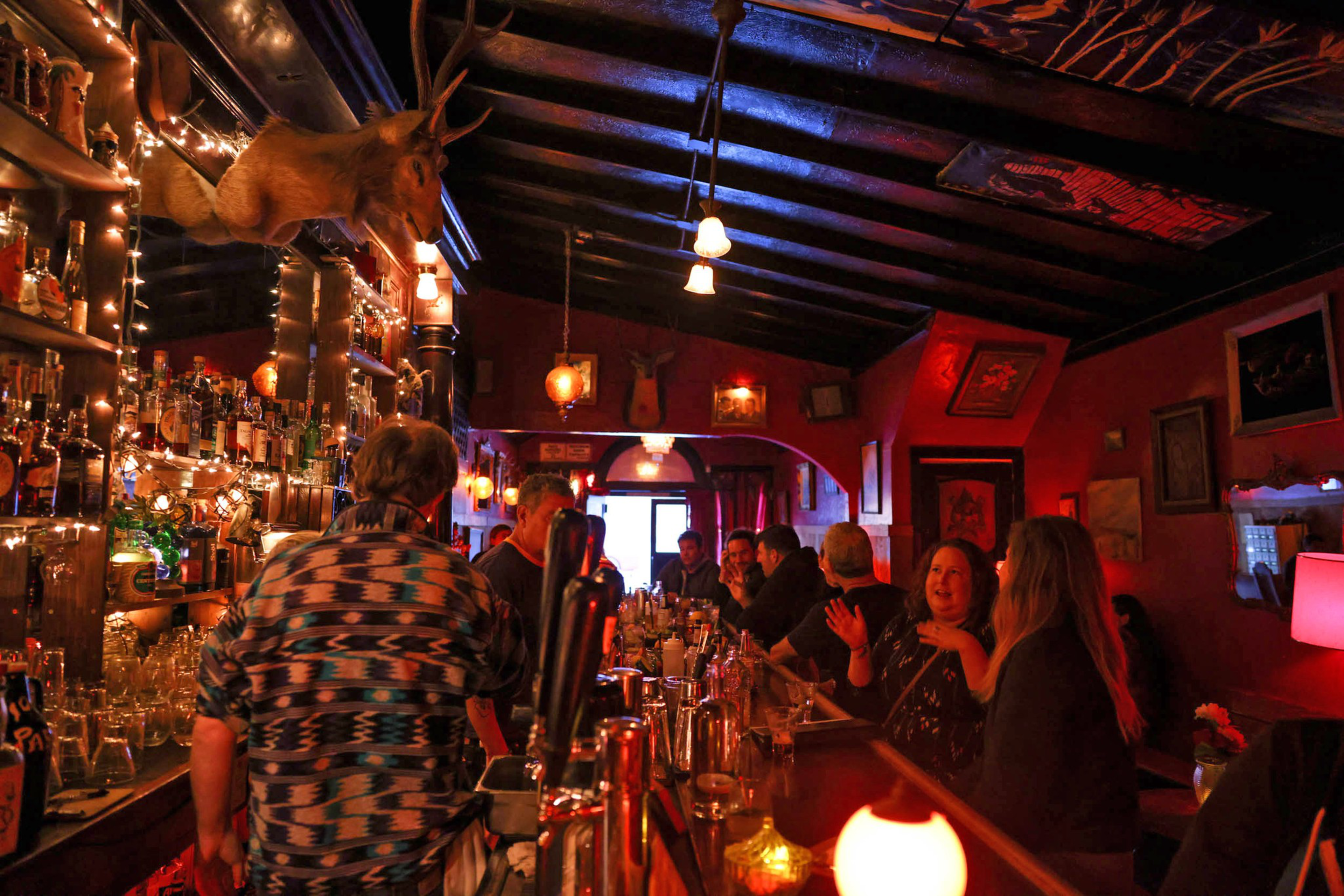 A dimly lit bar with red walls, filled with people chatting. Bottles line the bar shelves, and a mounted deer head is prominently displayed above.