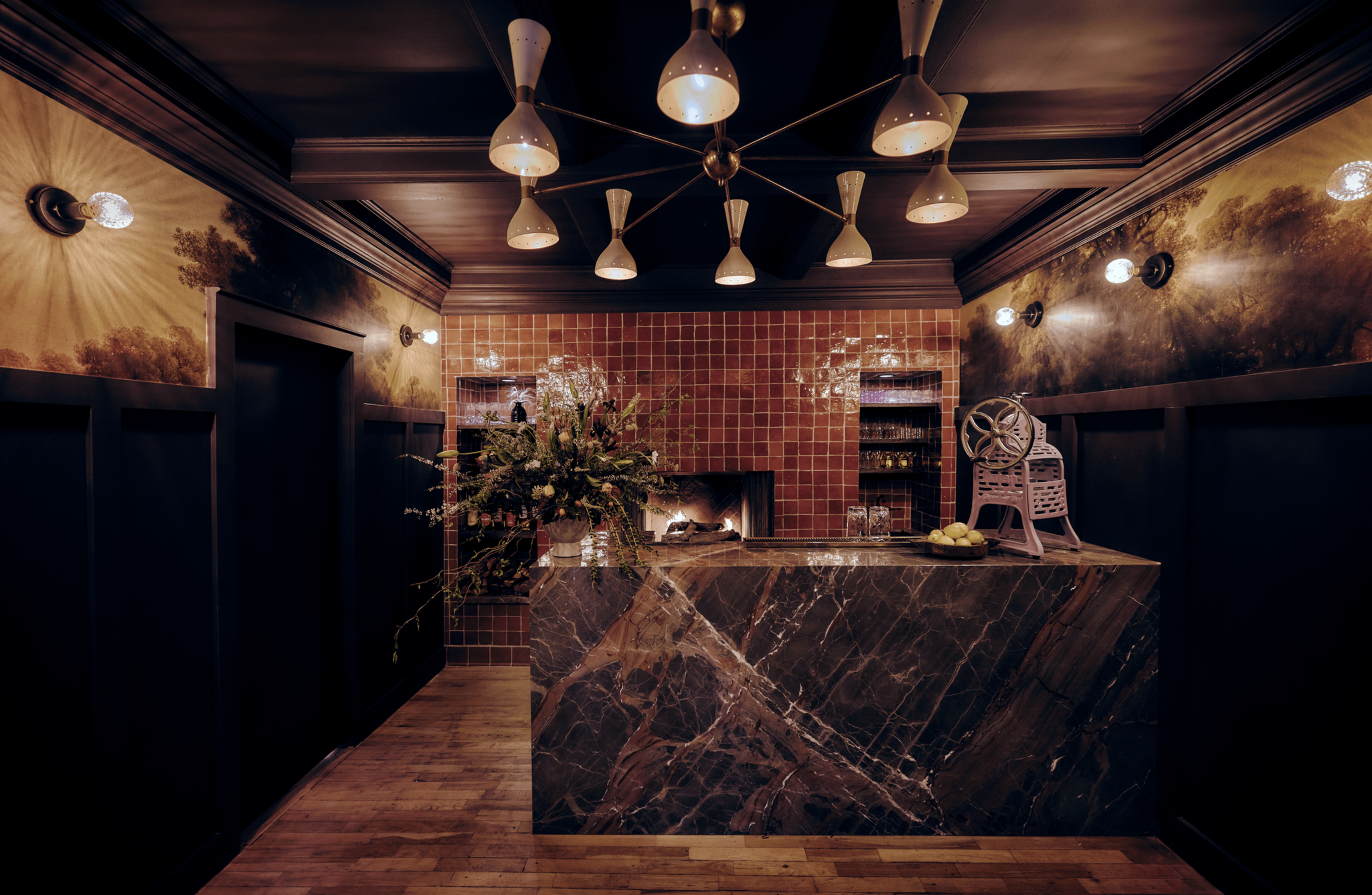 A stylish room with a marble bar, decorative lights, and a wall mural. A floral arrangement and an apple peeler sit on the bar in front of a tiled fireplace.