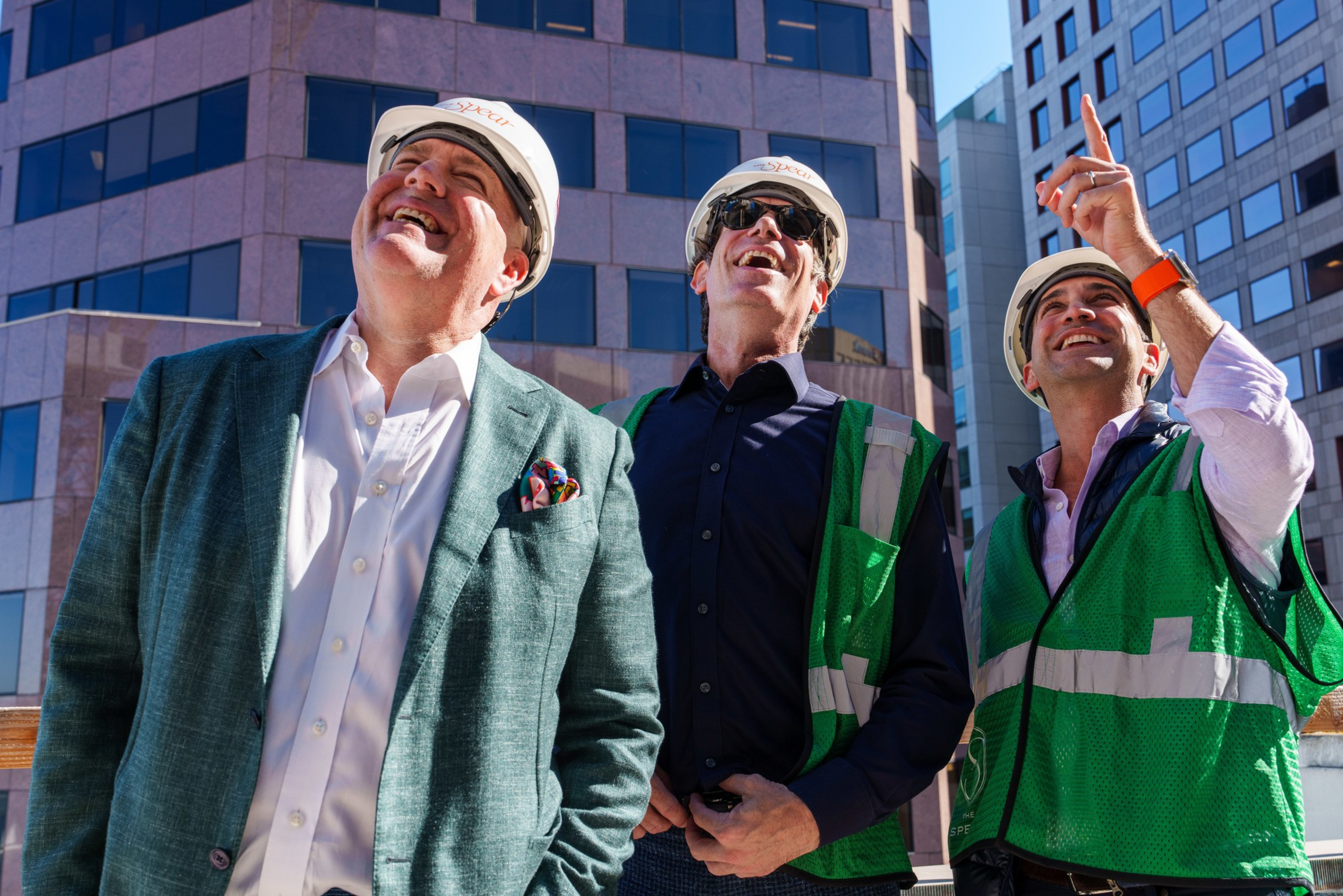 Three men wearing safety helmets and green vests stand outdoors in front of tall buildings, smiling and looking up, with one man pointing upwards.