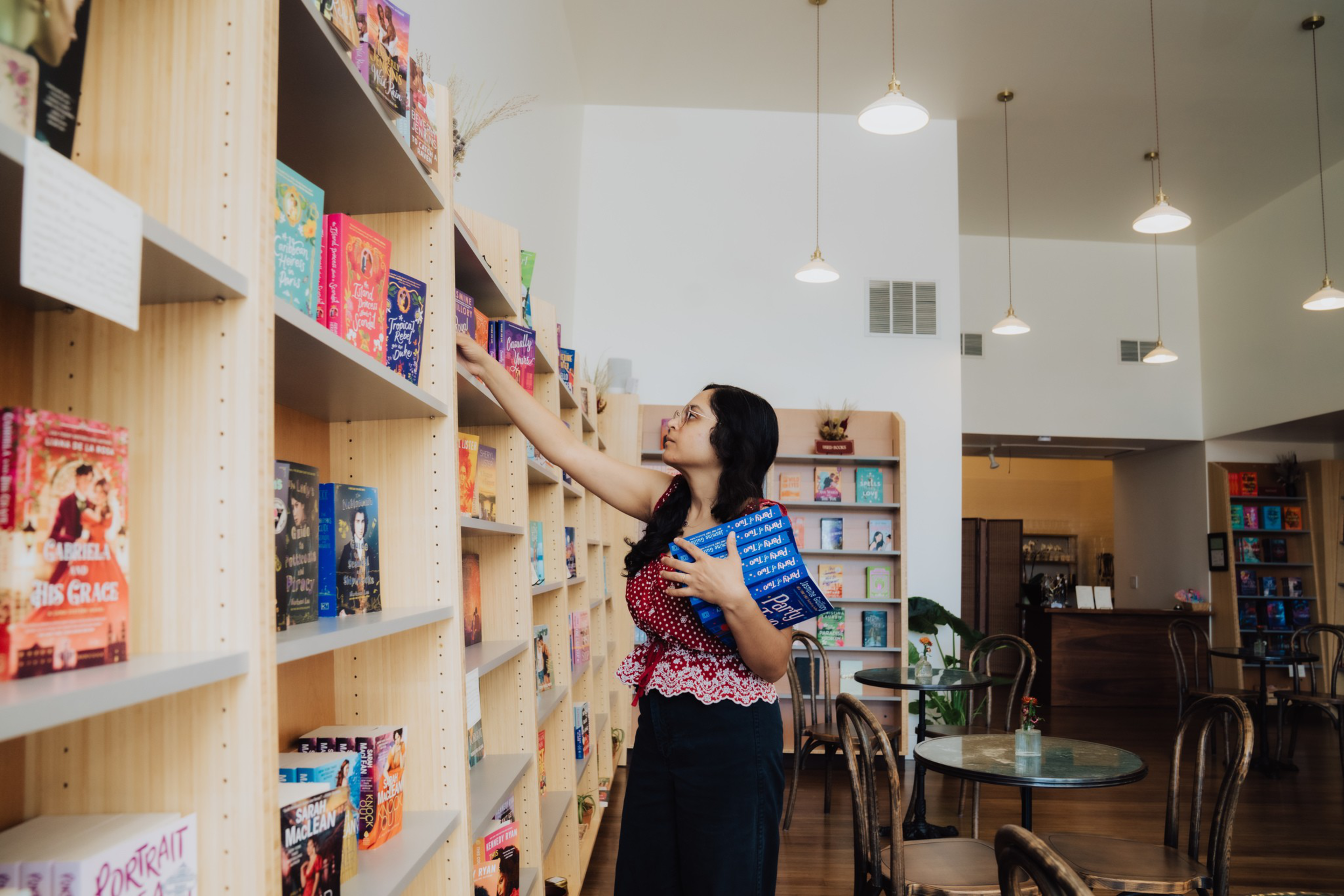 A woman in a red patterned top and black skirt reaches for a book on a tall wooden shelf in a bright, cozy bookstore or café.