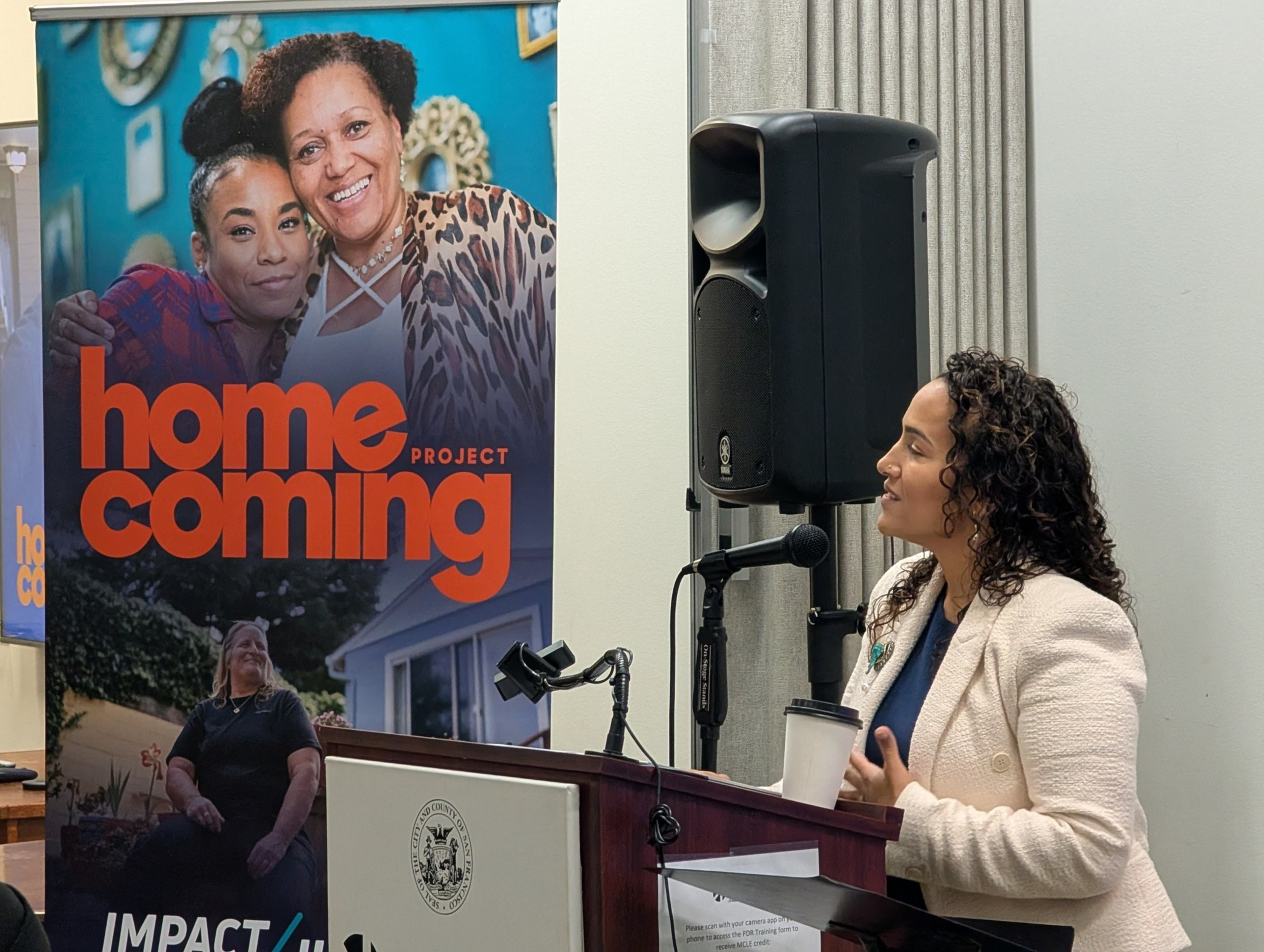 A woman with curly hair in a cream blazer speaks at a podium next to a large speaker and a “homecoming project” banner.