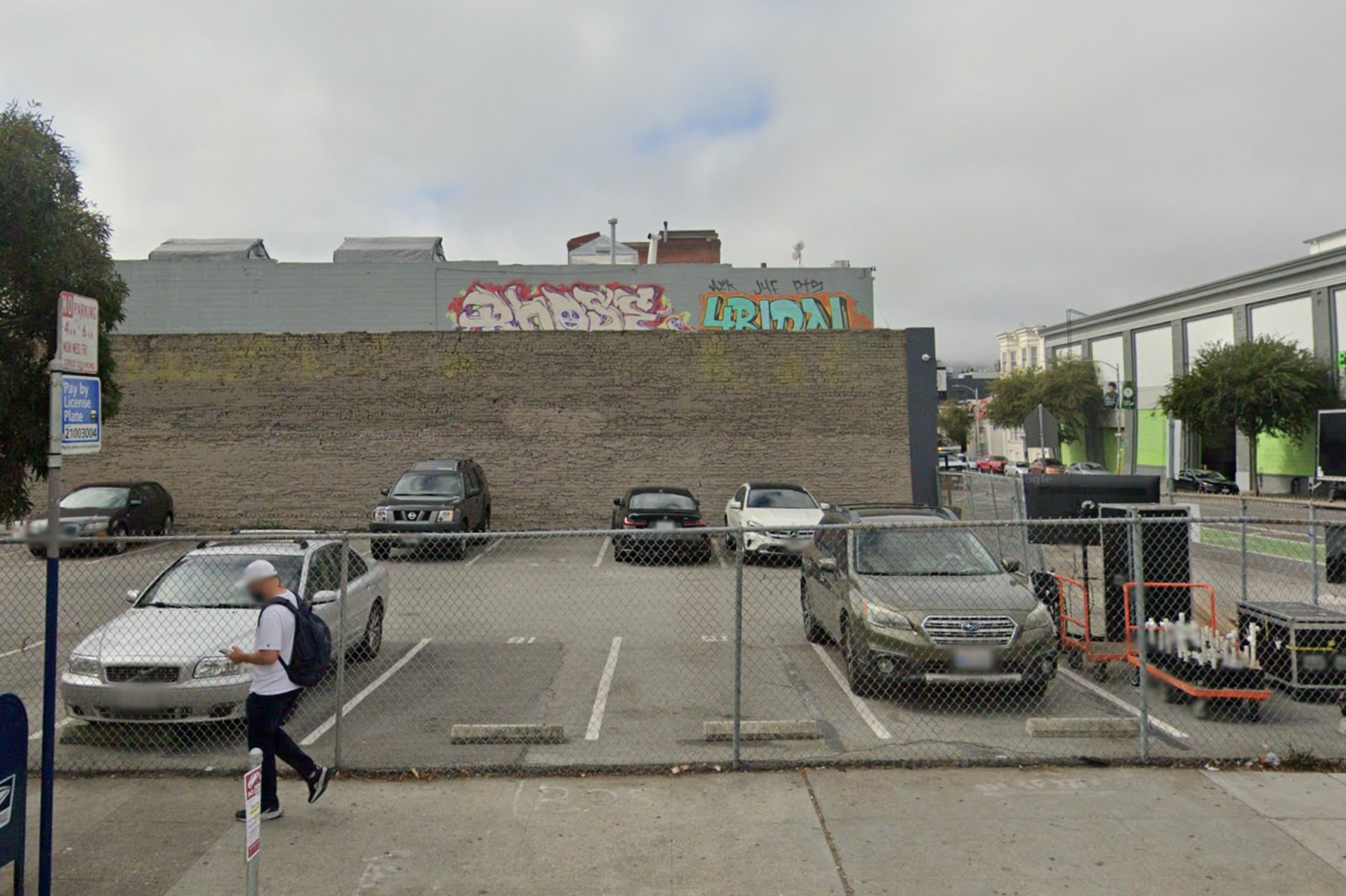 A small parking lot with five cars parked behind a chain-link fence and a person walking outside the fence on the sidewalk. Graffiti is on the wall behind.