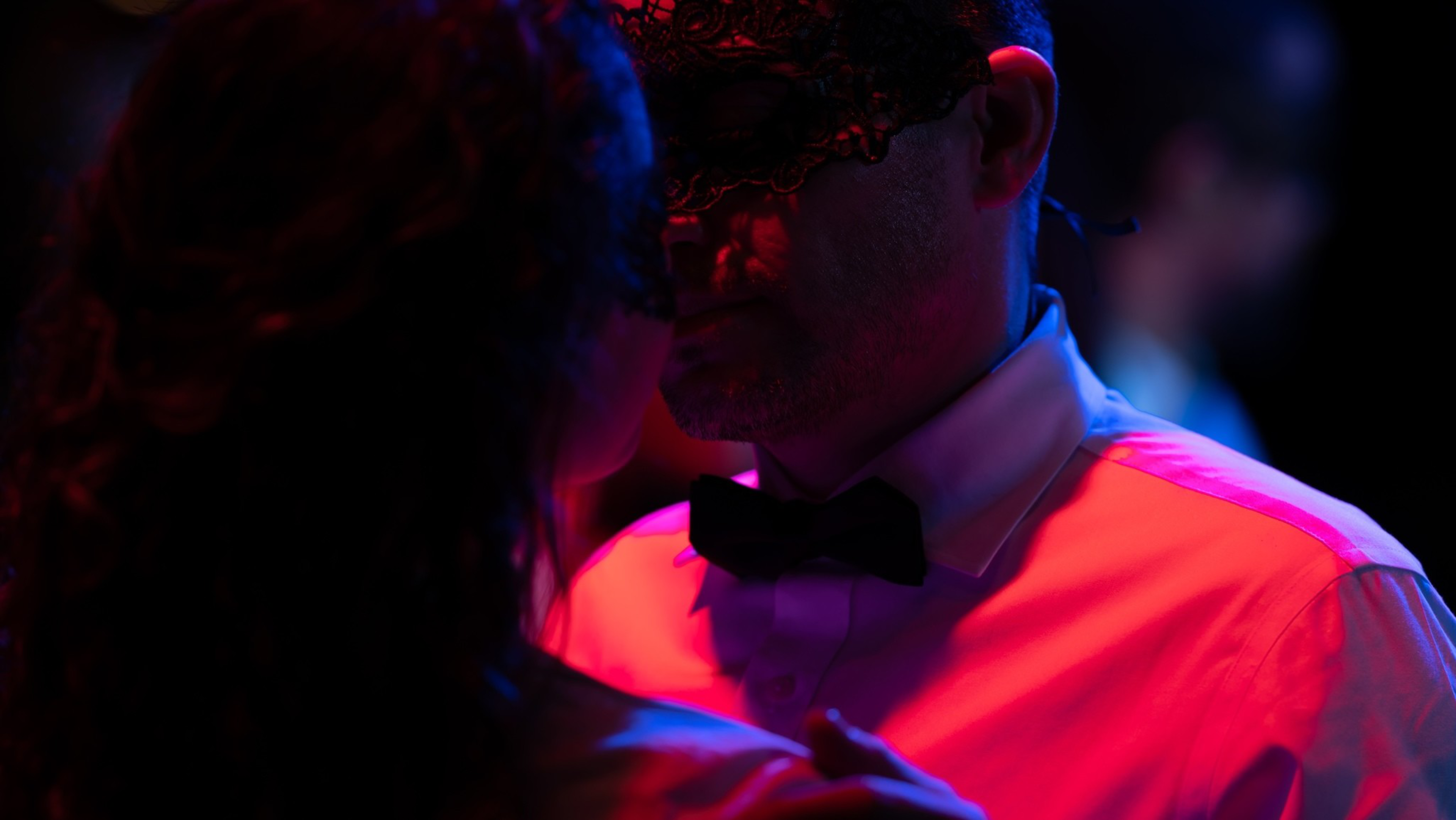 A couple wearing black lace masks dances closely under dim, colorful lighting, with the man dressed in a white shirt and black bow tie.