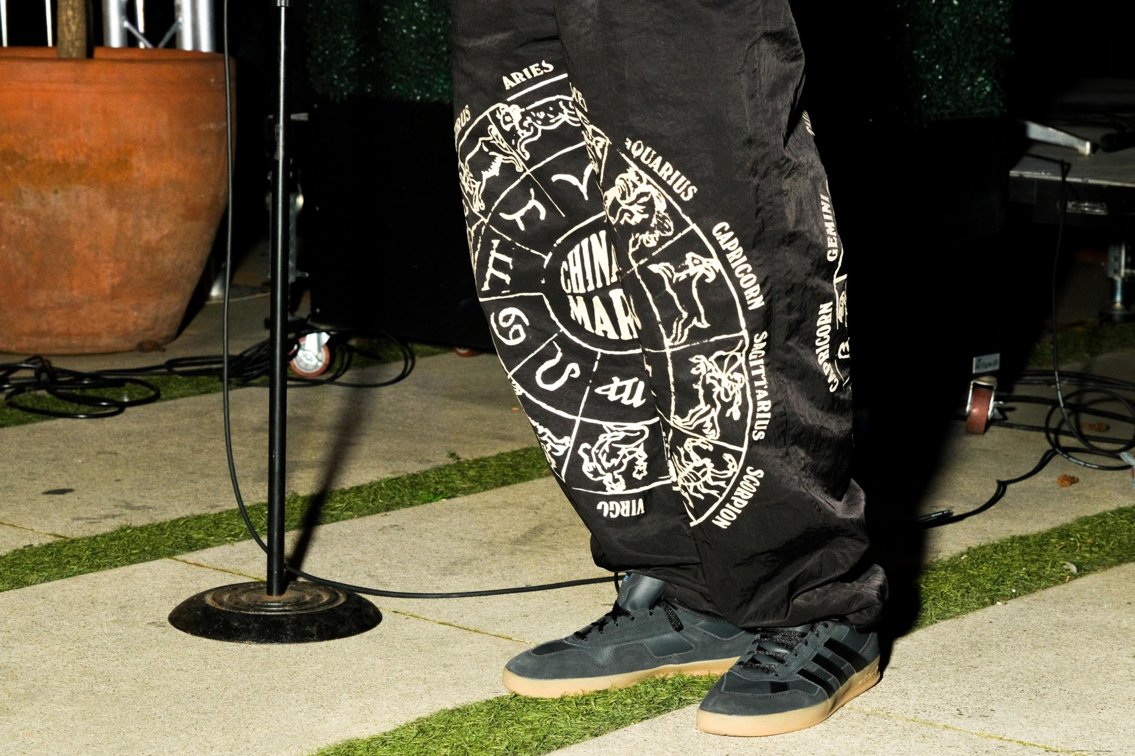 Black pants with zodiac signs and text, blue sneakers, standing near a mic stand on grass and concrete.