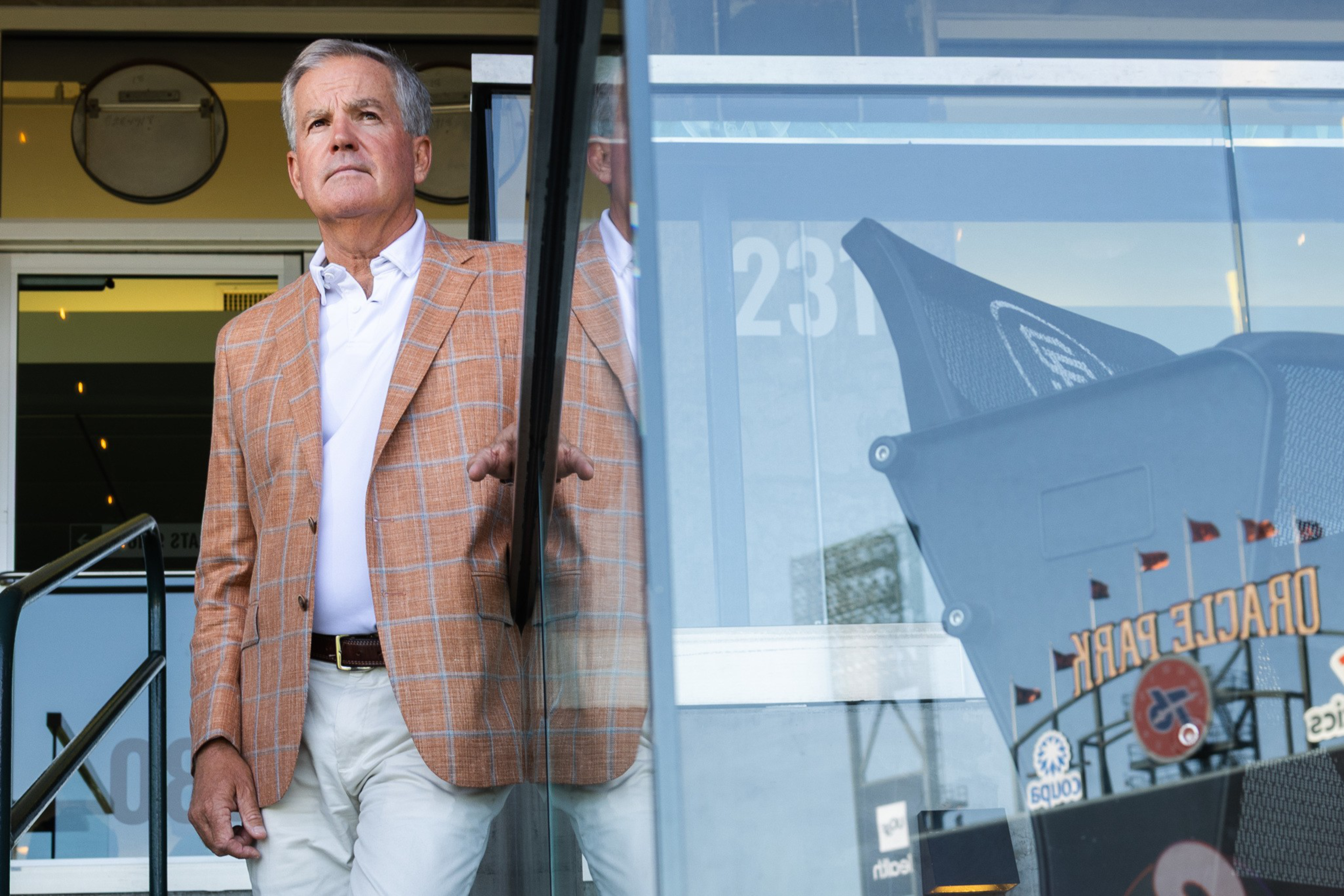 A man in a checkered orange blazer stands near a glass door with reflections, gazing outward. A sign reading &quot;Oracle Park&quot; is visible in the reflection.