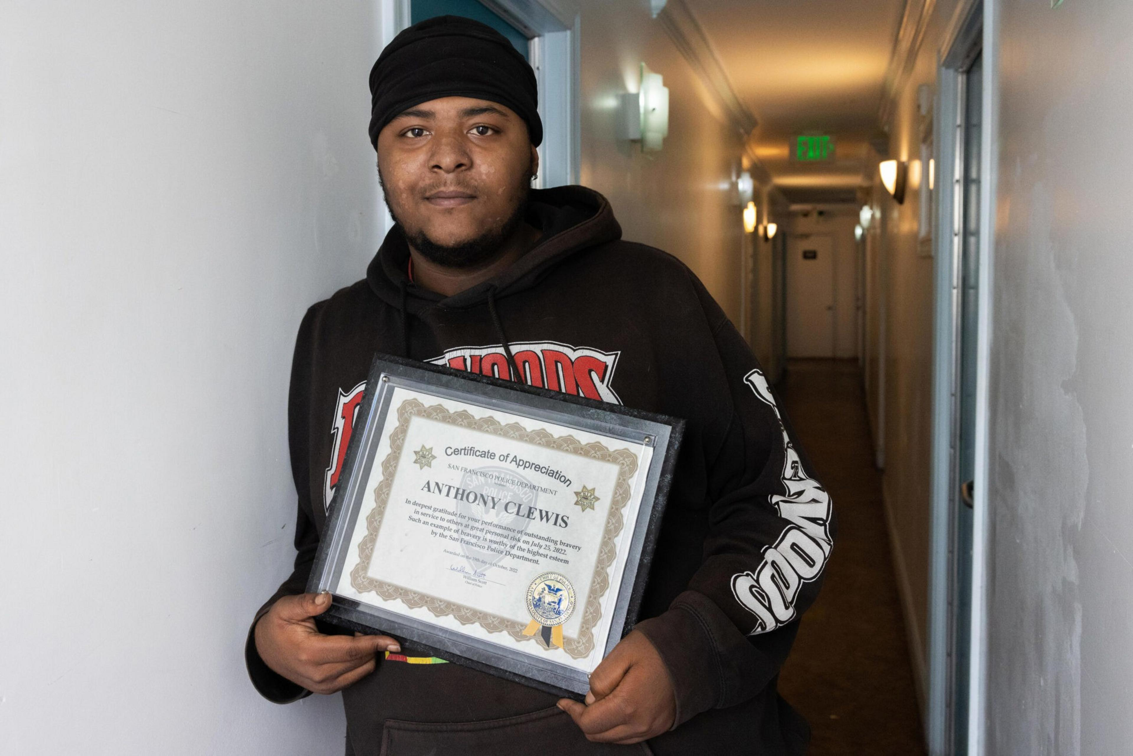 A man wearing a black beanie and hoodie stands in a hallway, holding a framed certificate of appreciation with the name Anthony Clewis.
