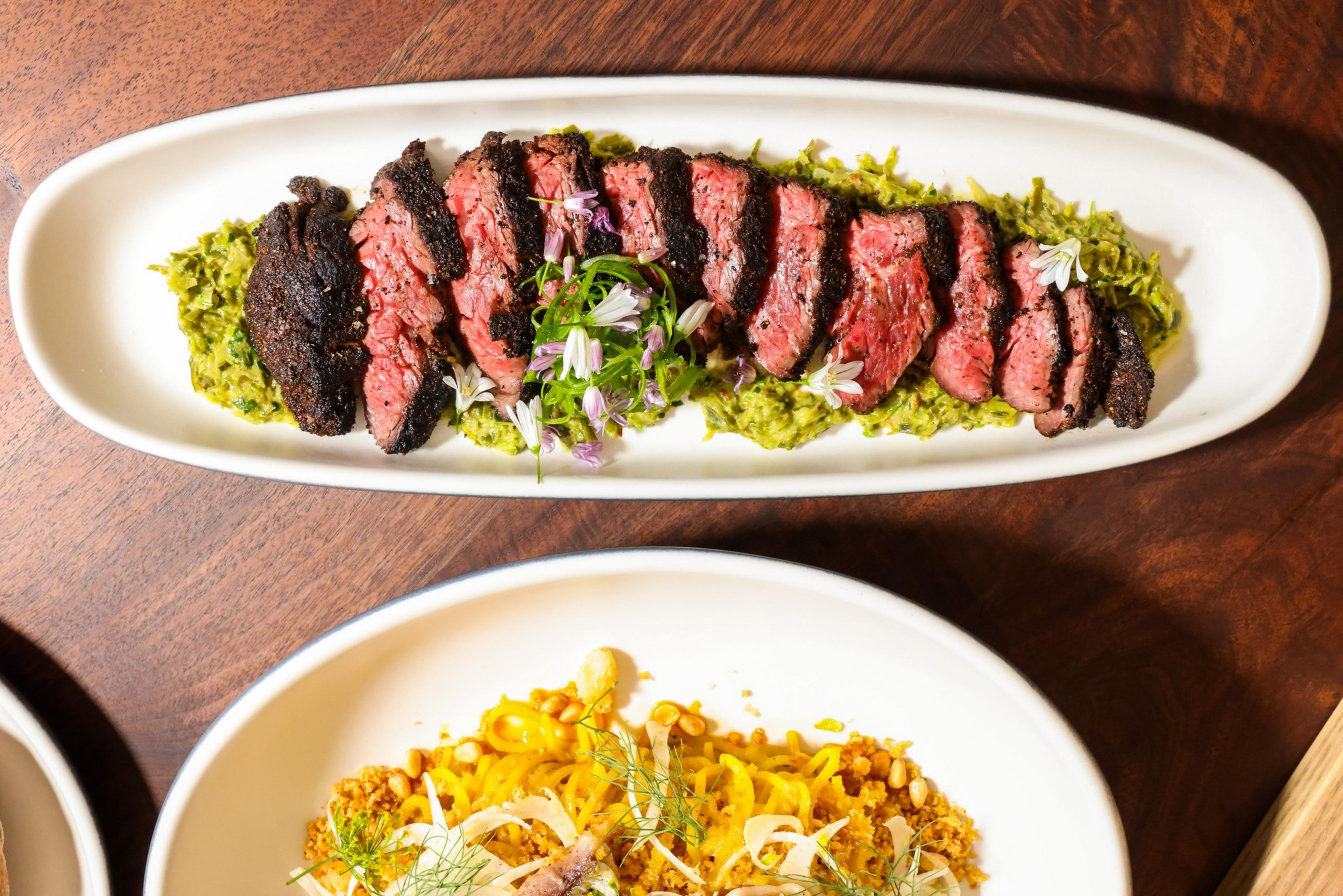 Slices of medium-rare steak are arranged on a green sauce, garnished with white flowers and herbs on a white oval plate.