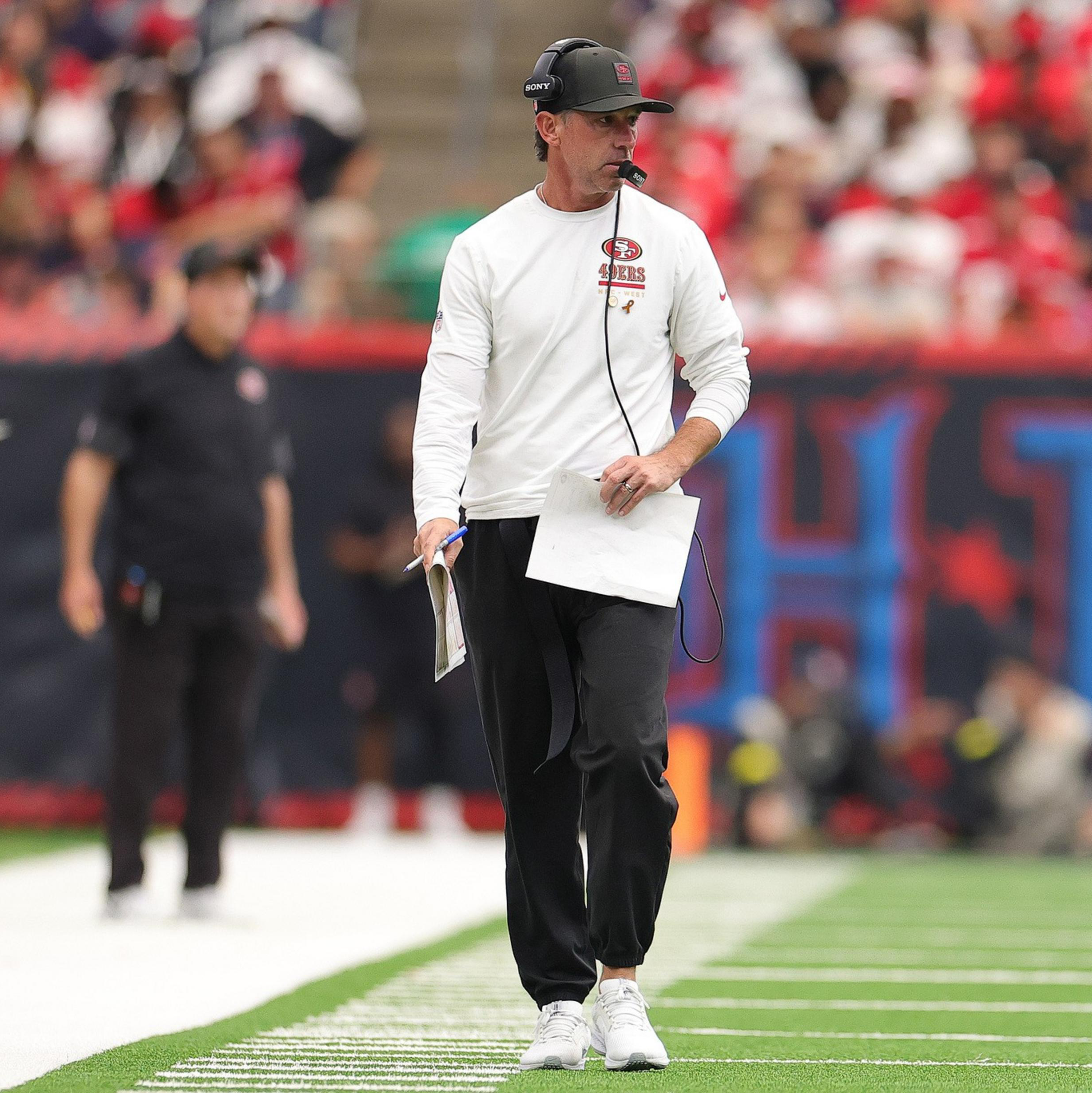 A football coach in a white 49ers shirt, black pants, and headset walks on the sideline holding papers during a game with blurred crowd in the background.