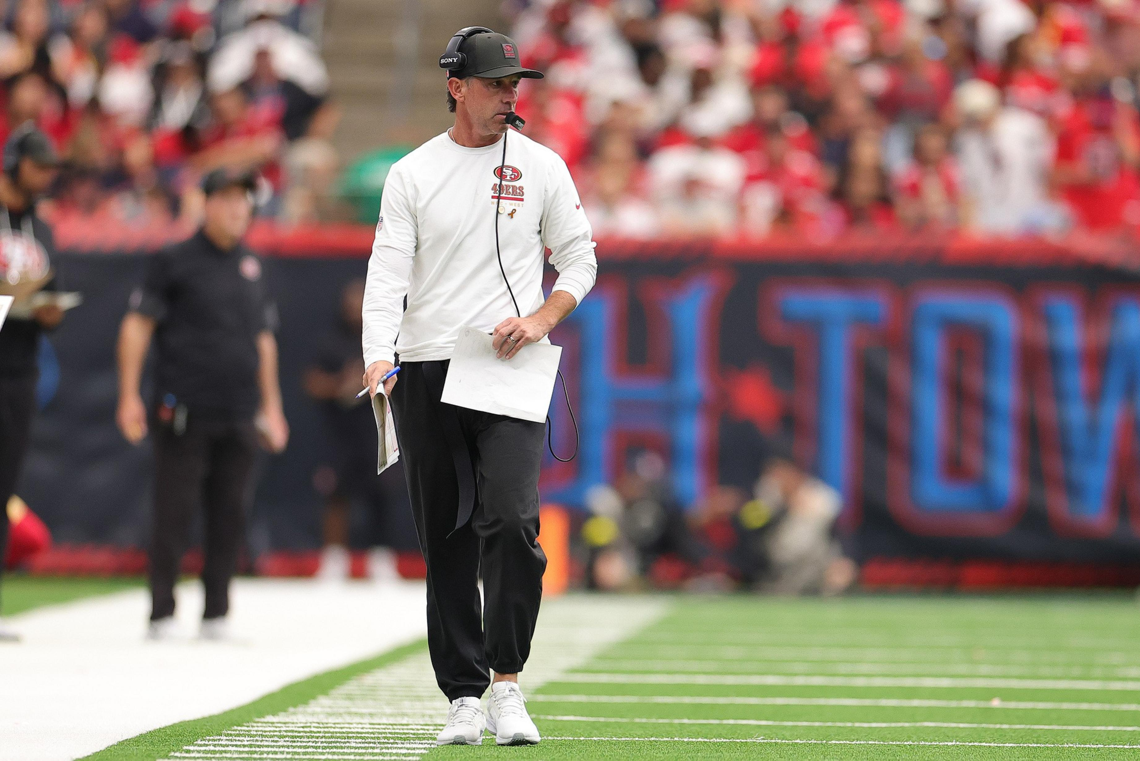 A football coach in a white 49ers shirt, black pants, and headset walks on the sideline holding papers during a game with blurred crowd in the background.