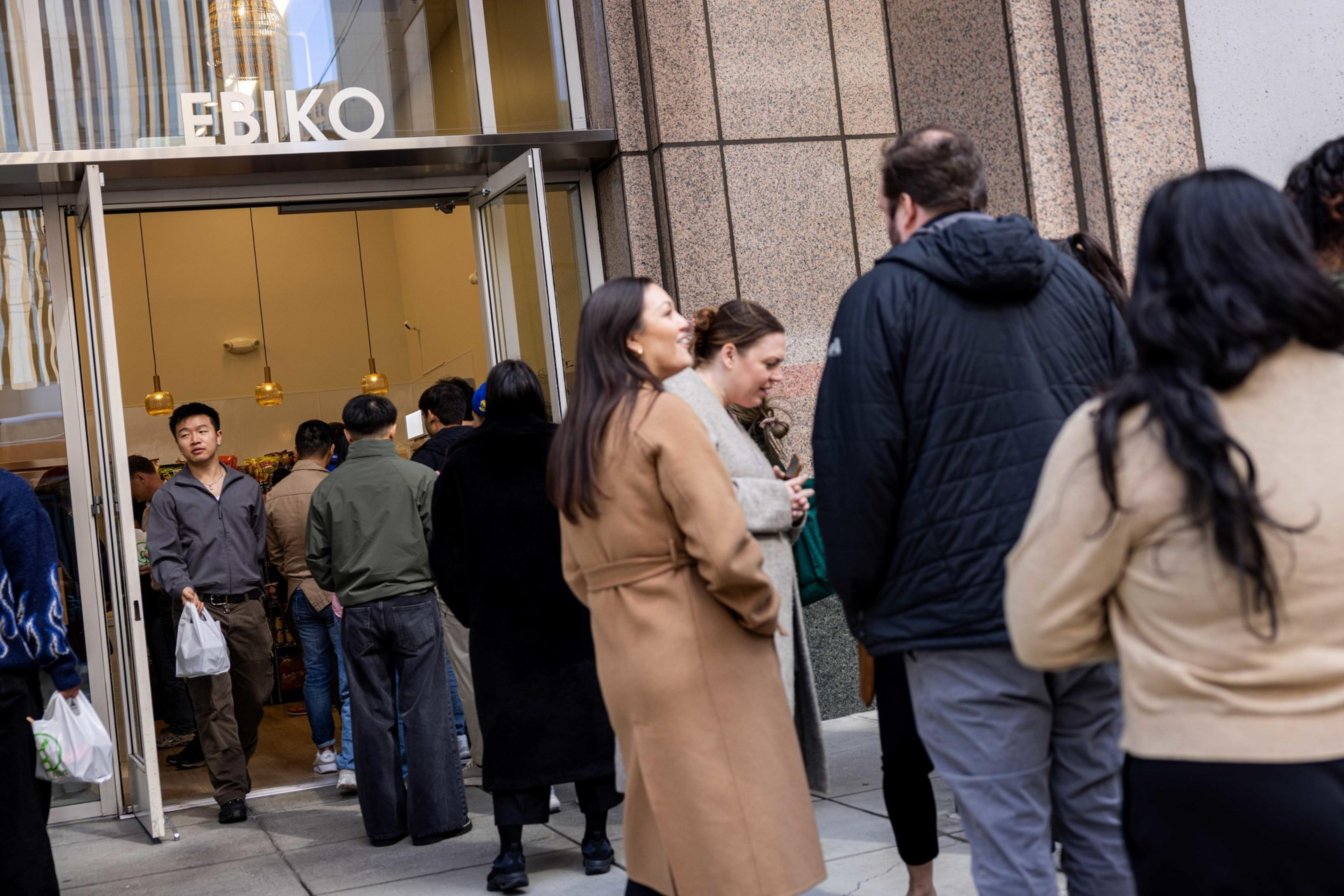 A group of people is waiting in line outside a store named &quot;EBIKO.&quot; The entrance is open, and people are entering, with some carrying bags.