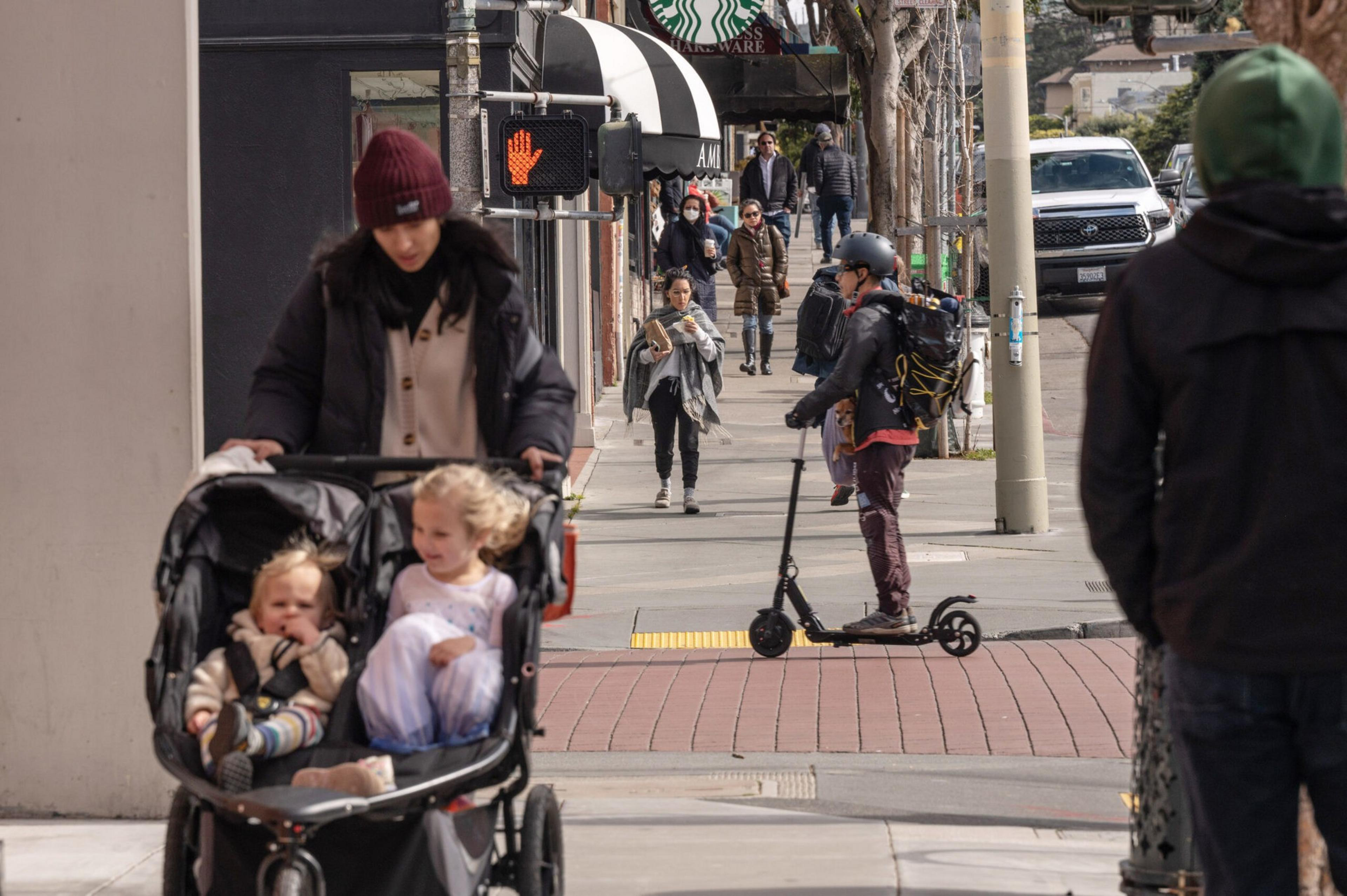 A woman pushes a double stroller with two children while a person rides an electric scooter across a city sidewalk with several pedestrians walking.