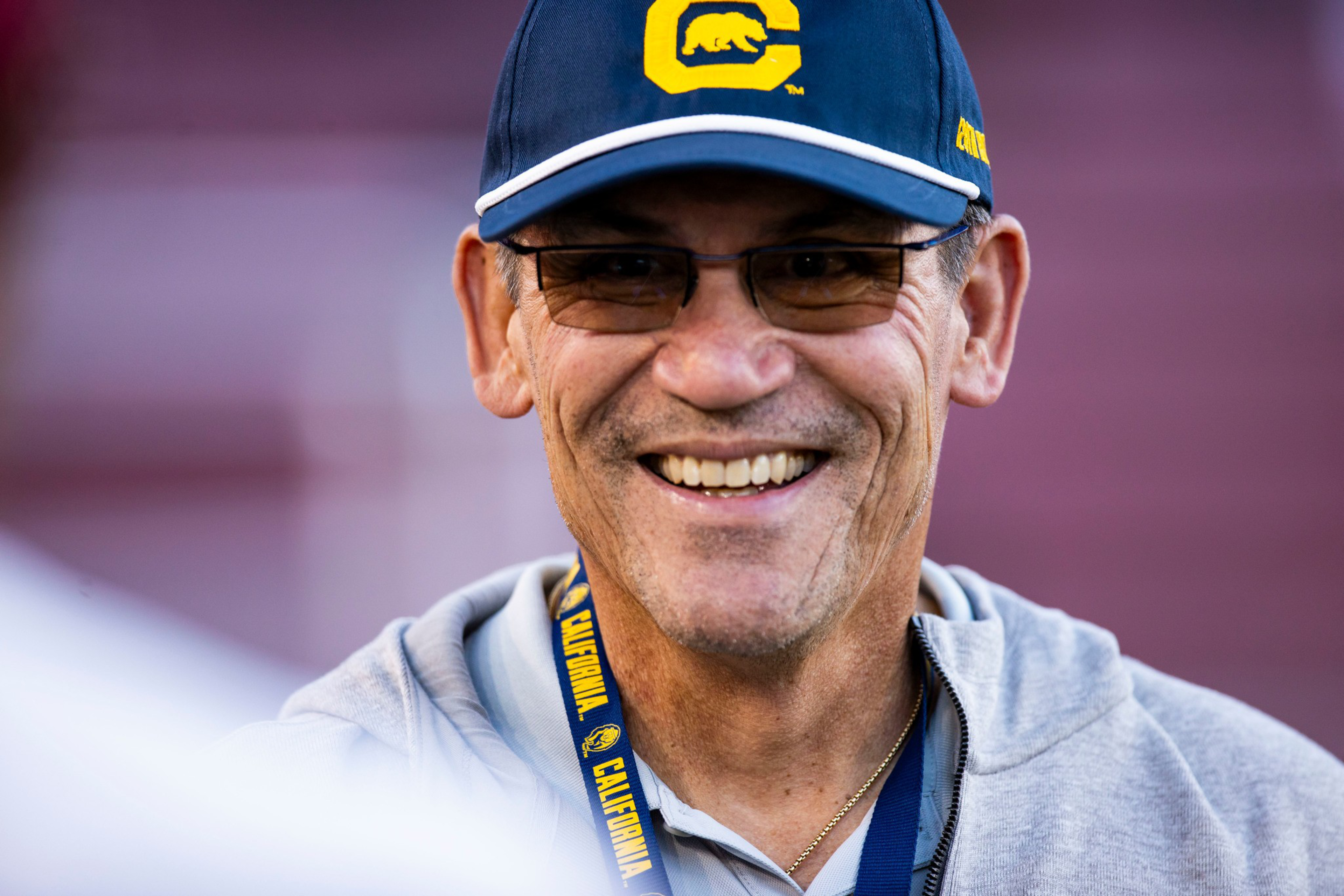 A man wearing glasses, a blue cap with a yellow "C" and bear logo, a gray hoodie, and a lanyard that says "Cal" and "California" smiles warmly.