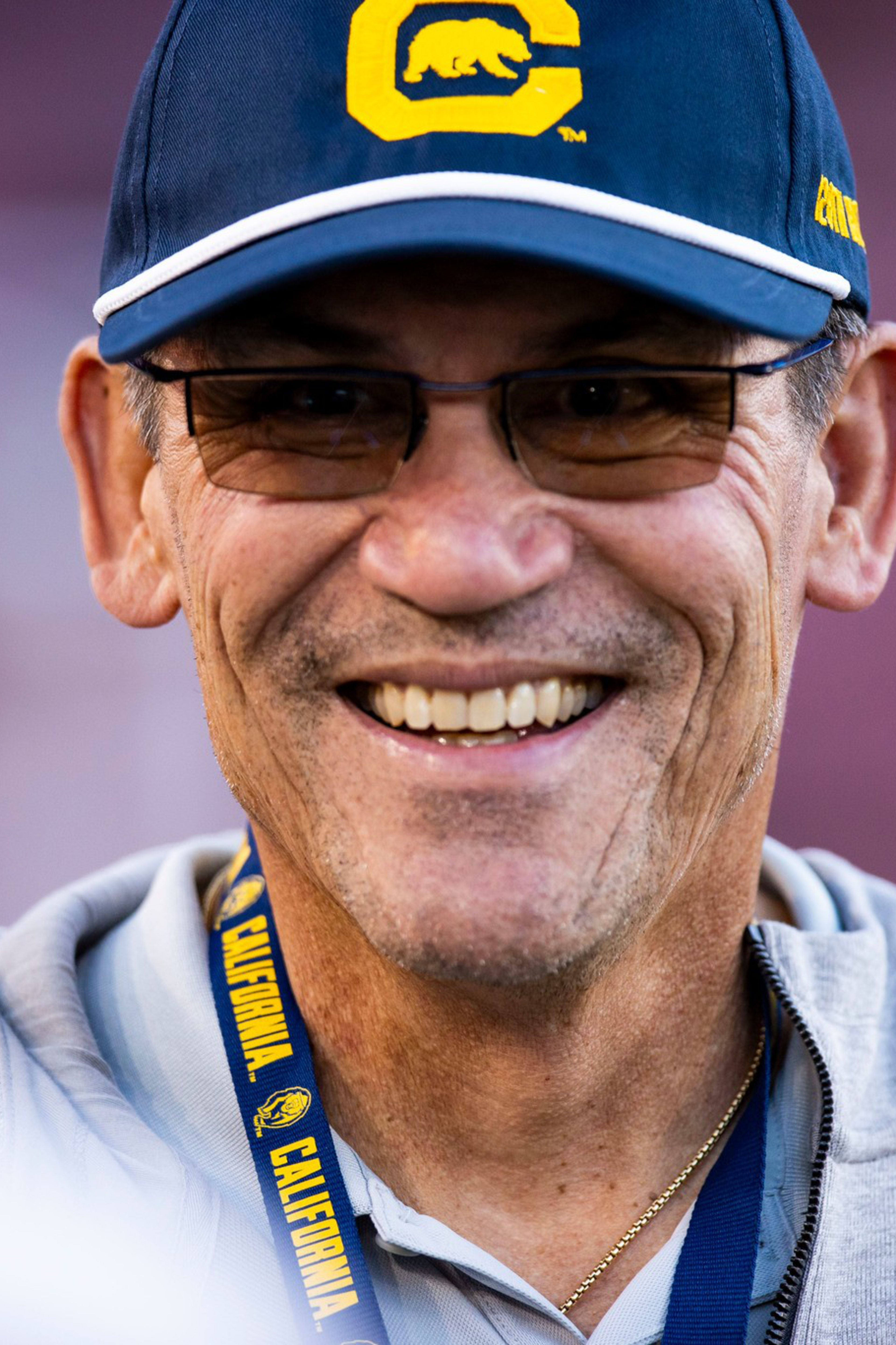 A man wearing glasses, a blue cap with a yellow "C" and bear logo, a gray hoodie, and a lanyard that says "Cal" and "California" smiles warmly.