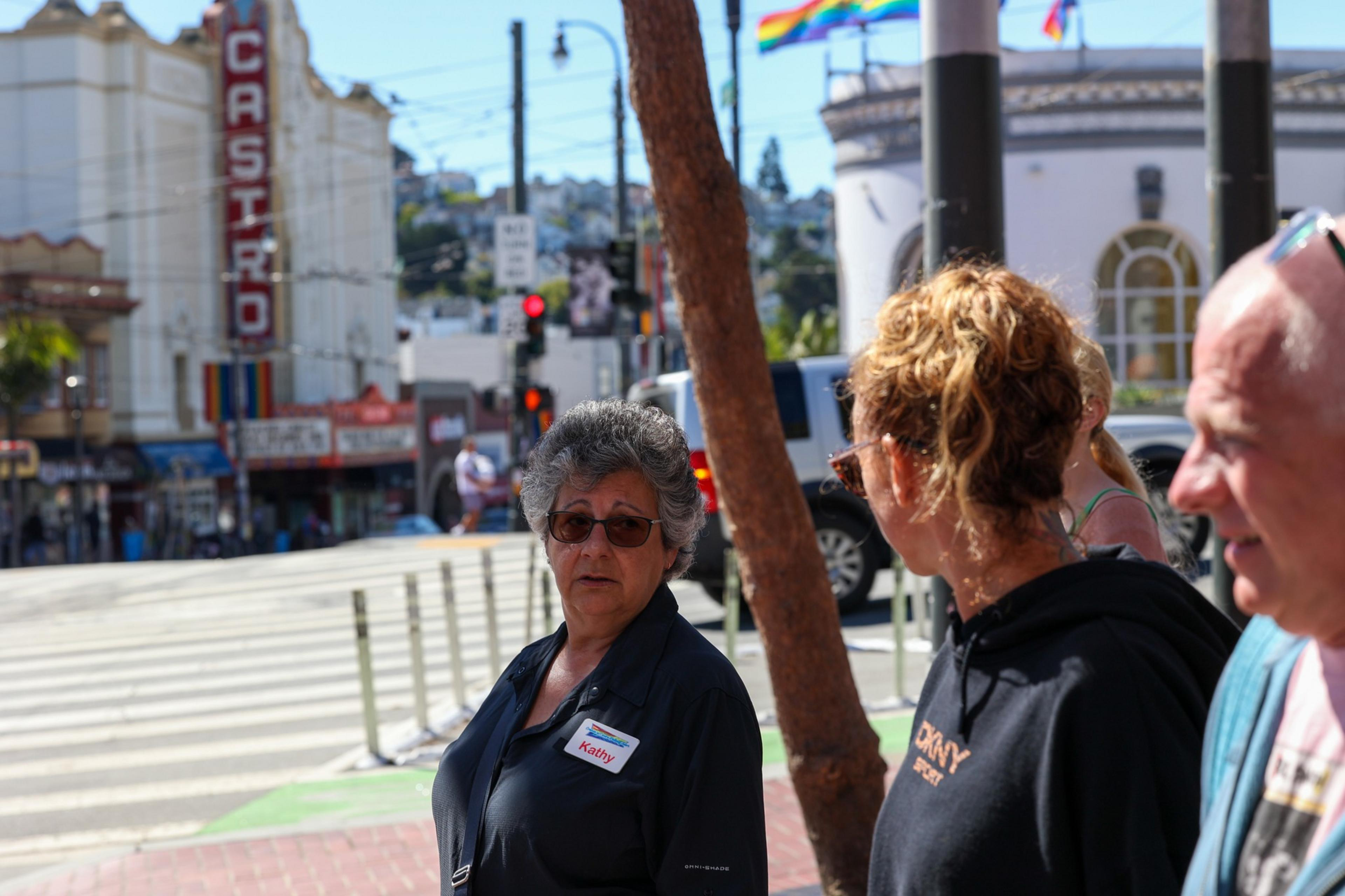 An older woman wearing glasses and a name tag stands near a crosswalk while two people talk, with a Castro Theatre sign and pride flags in the background.